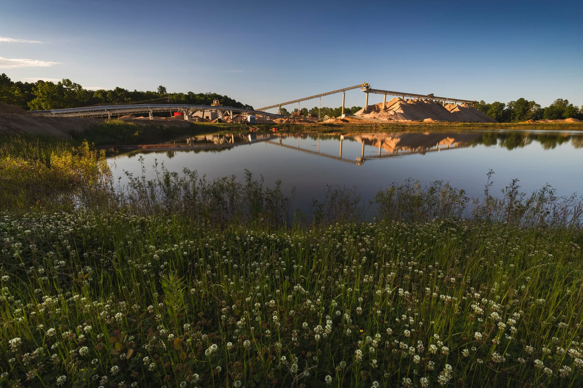 A body of water reflecting a large gravel plant with conveyor belts and piles of gravel in the background, surrounded by green vegetation and flowers in the foreground.