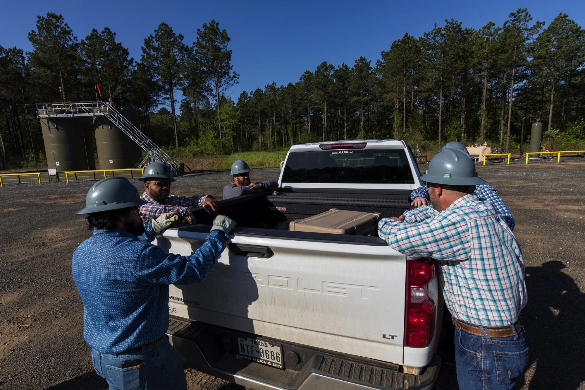 Six workers wearing safety helmets and gloves are loading equipment into the bed of a white pickup truck on a construction site with trees and industrial tanks in the background.