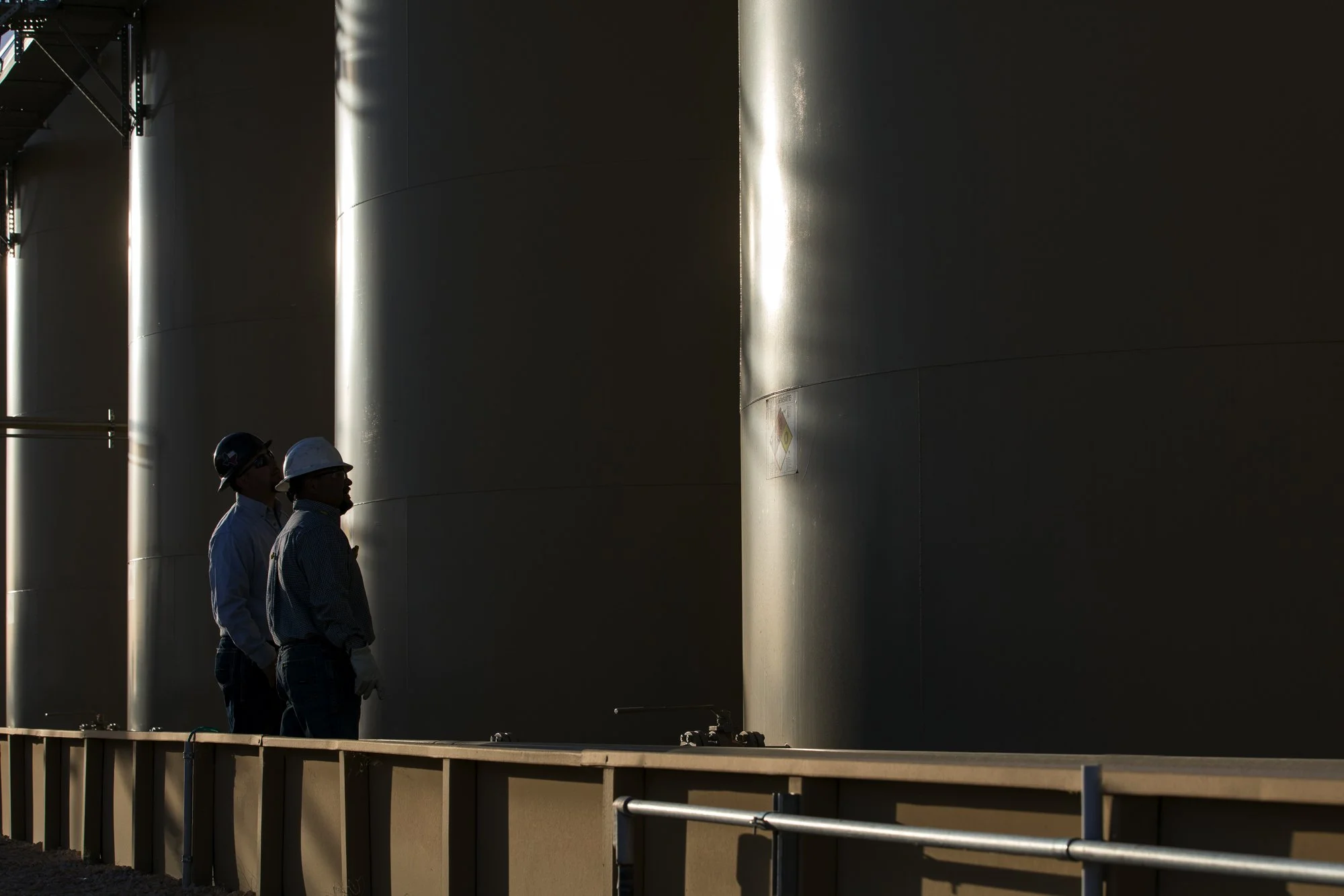 Two workers in safety helmets and work attire observing large industrial tanks.