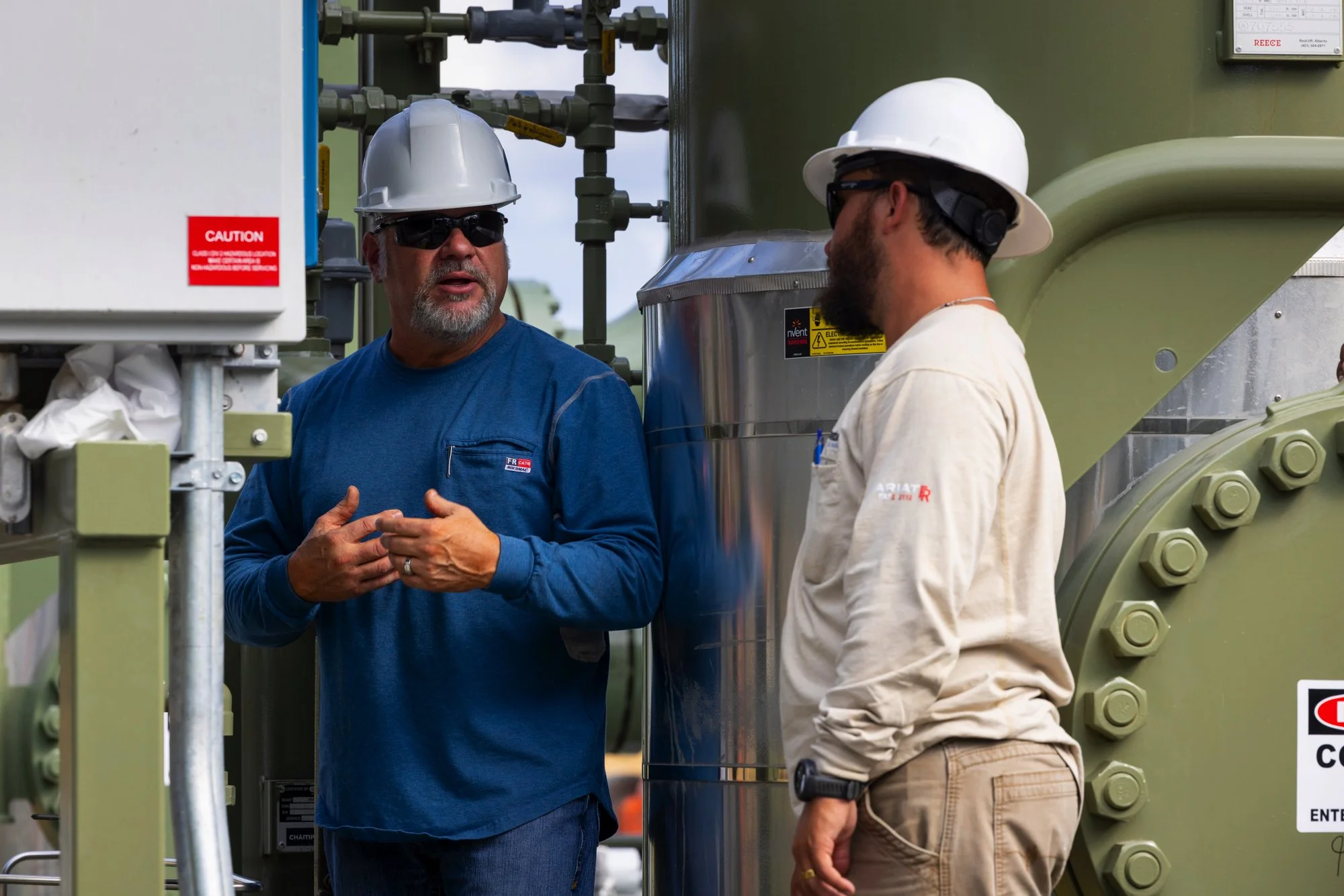 Two men wearing hard hats and safety glasses standing and talking near industrial machinery outdoors.