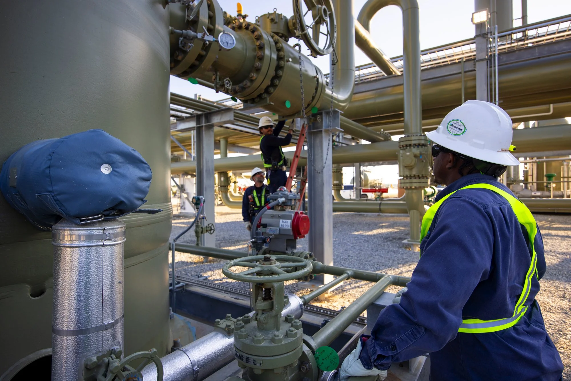 Workers in safety gear and helmets inspecting or maintaining large industrial pipes and equipment at an industrial facility, possibly an oil or gas plant.