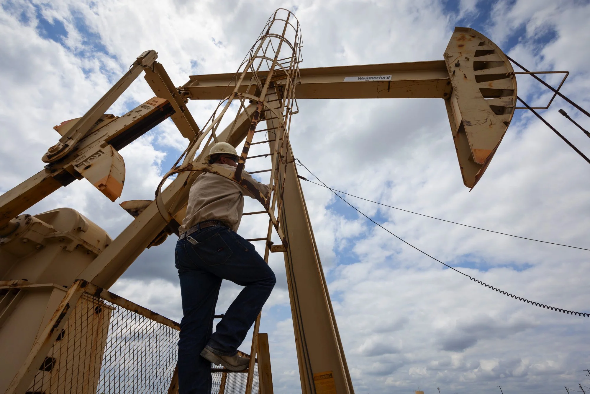 A worker wearing a helmet climbing a ladder on an oil pumpjack against a cloudy sky.