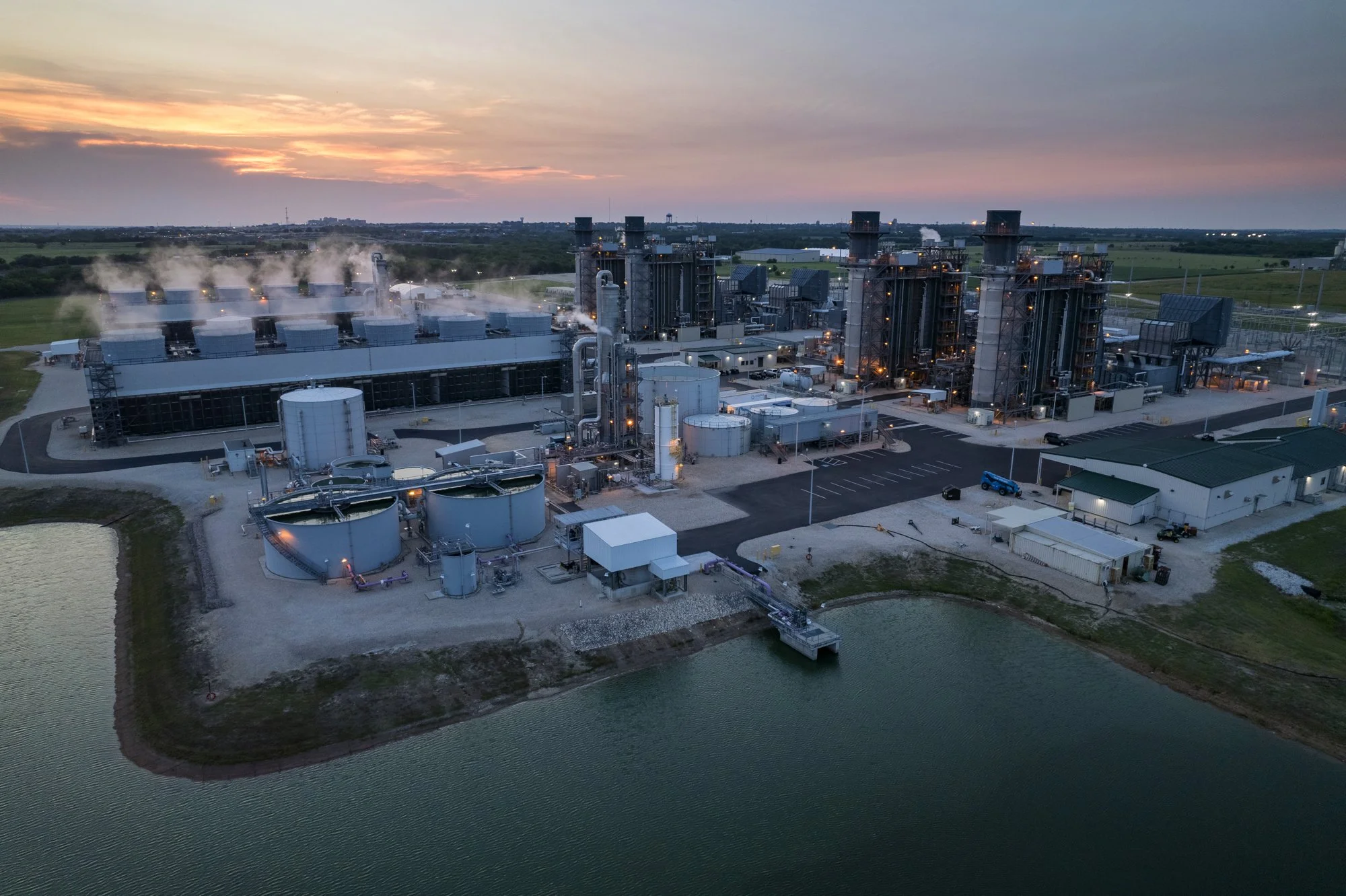 Aerial view of an industrial complex near a body of water at sunset, with large storage tanks, multiple buildings, and chimneys emitting steam or smoke.