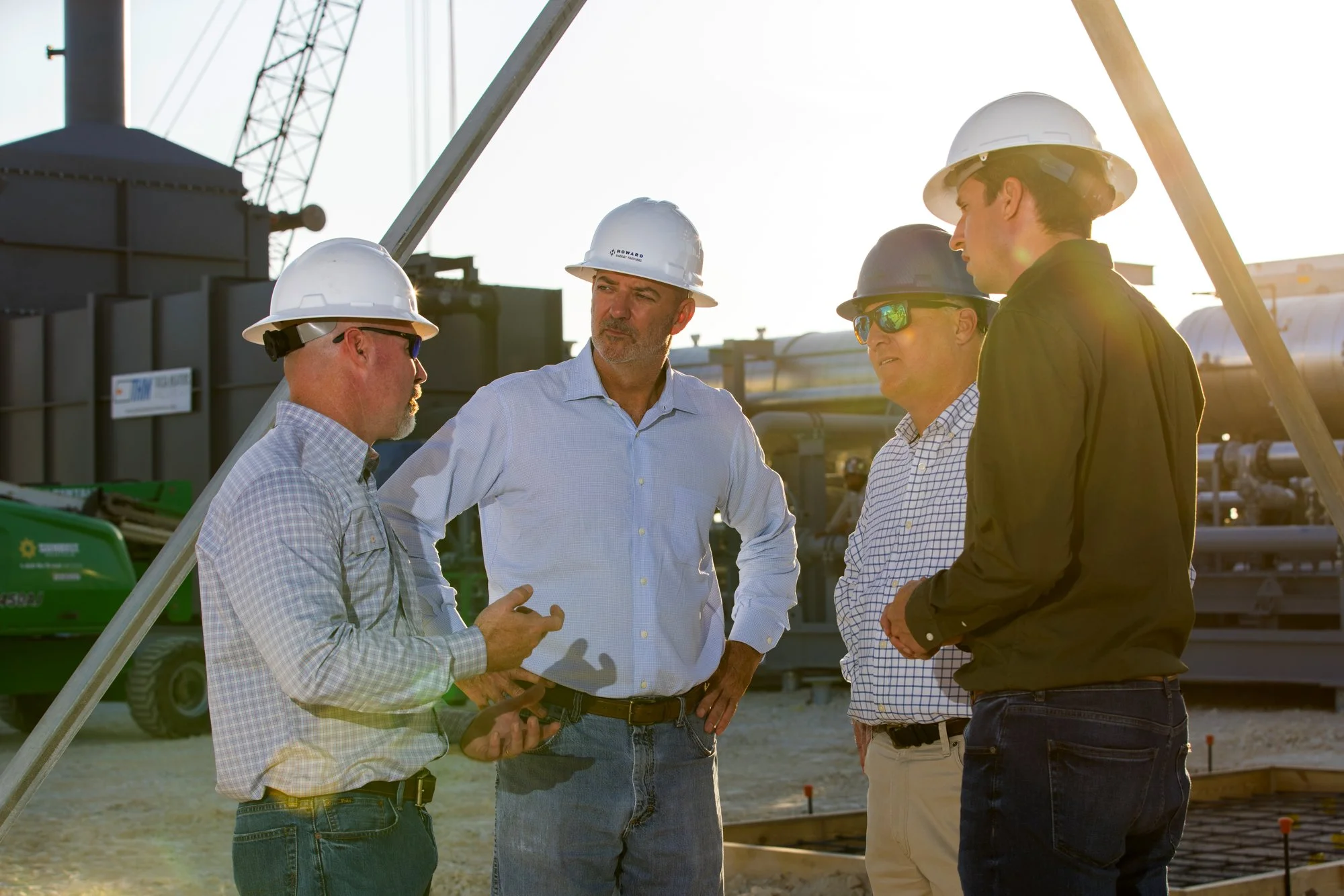 Four men wearing safety helmets and casual business attire having a discussion at a construction site with industrial equipment in the background.