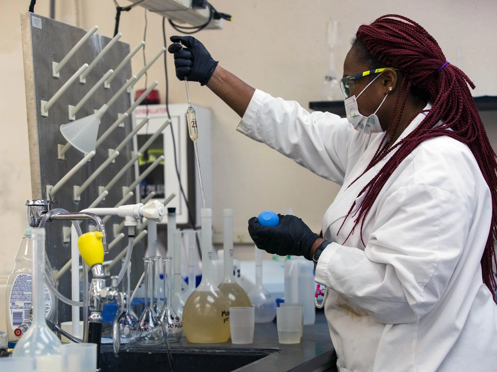 A scientist wearing a white lab coat, face mask, and safety glasses conducts a chemistry experiment in a laboratory, using a pipette and various glassware.