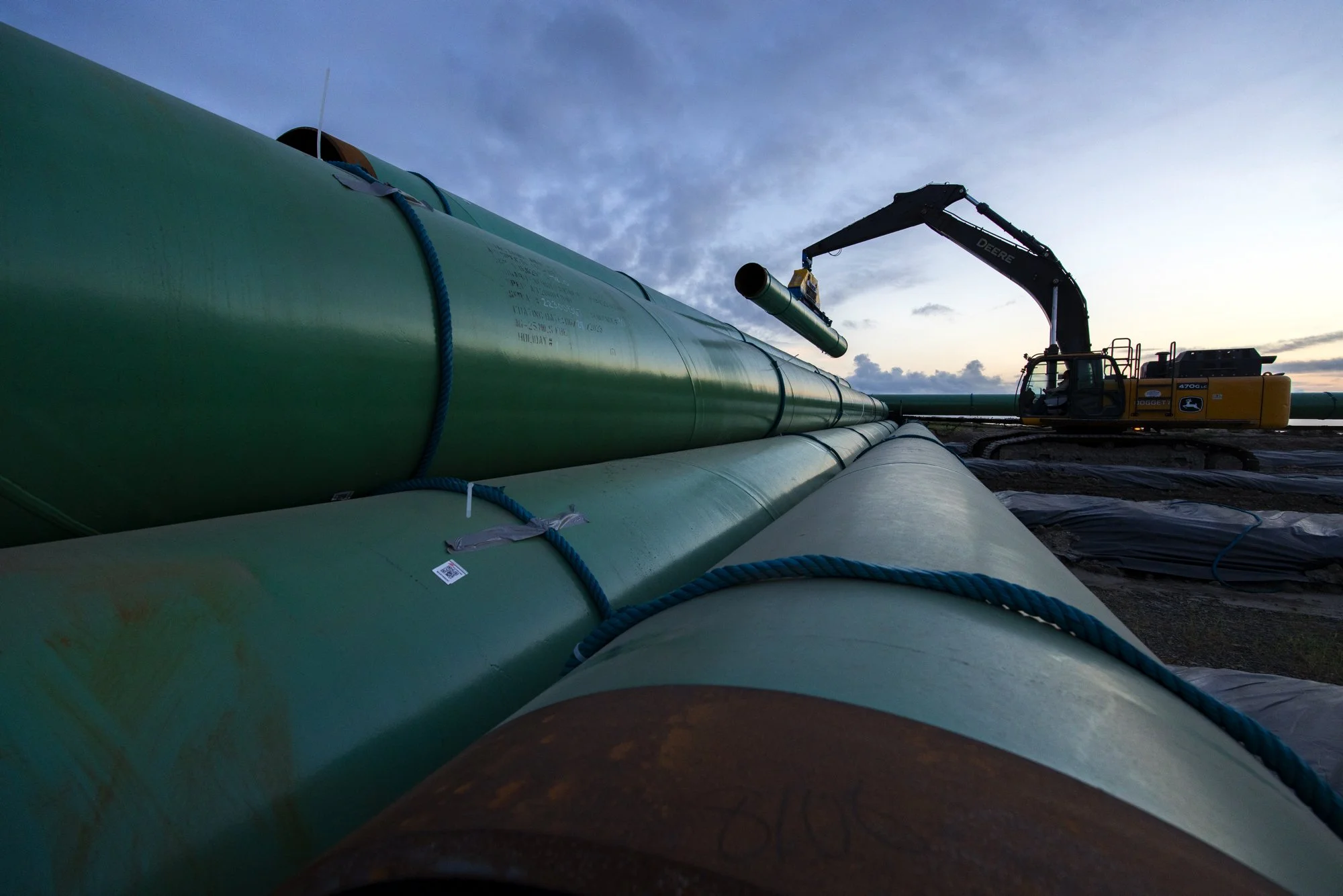 Large green oil pipeline under construction, with a crane lifting a segment into place at sunset.