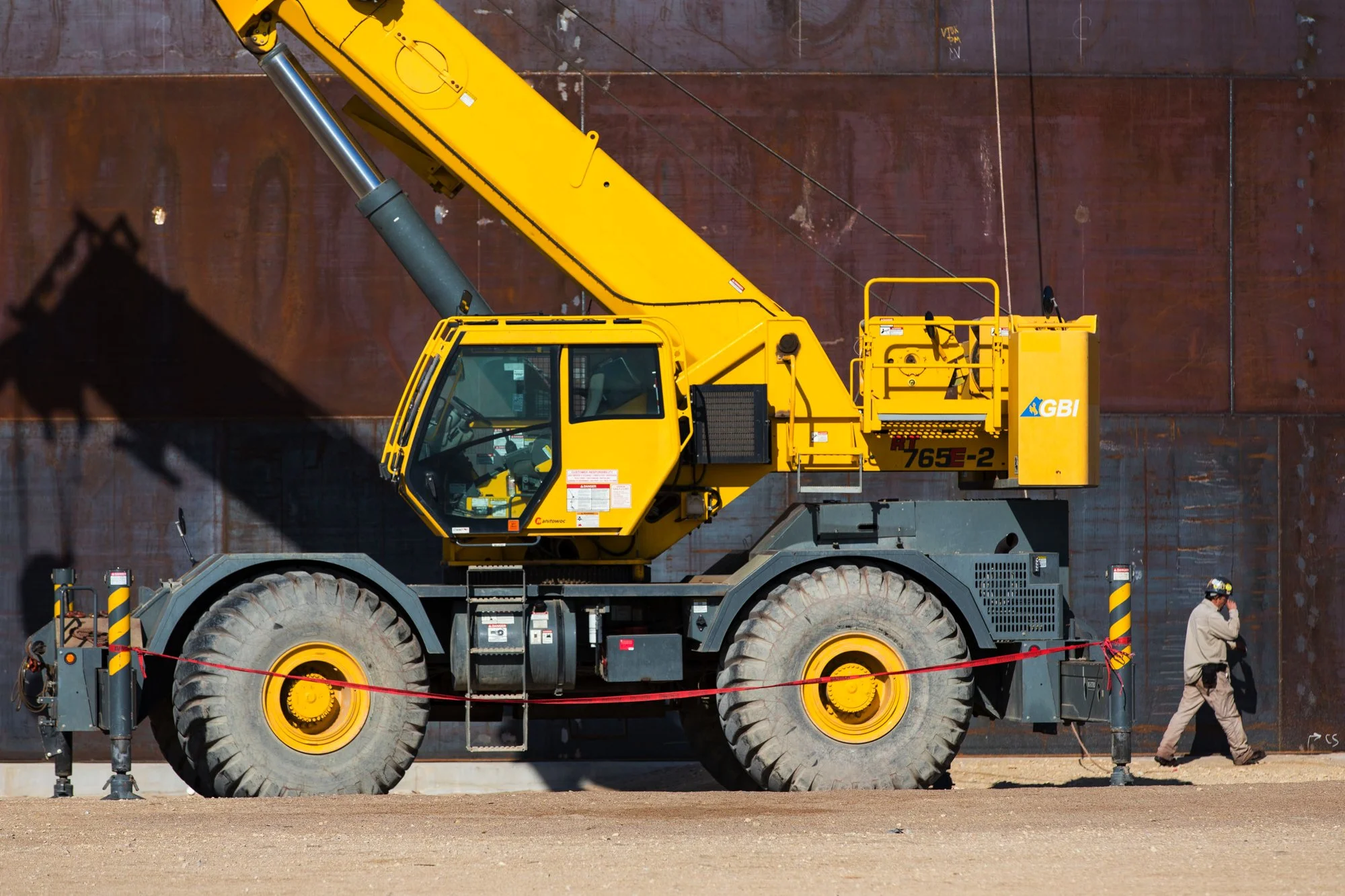 A large yellow crane with black wheels and a long arm is positioned next to a rusted metal wall, with a worker walking and talking on the phone nearby.