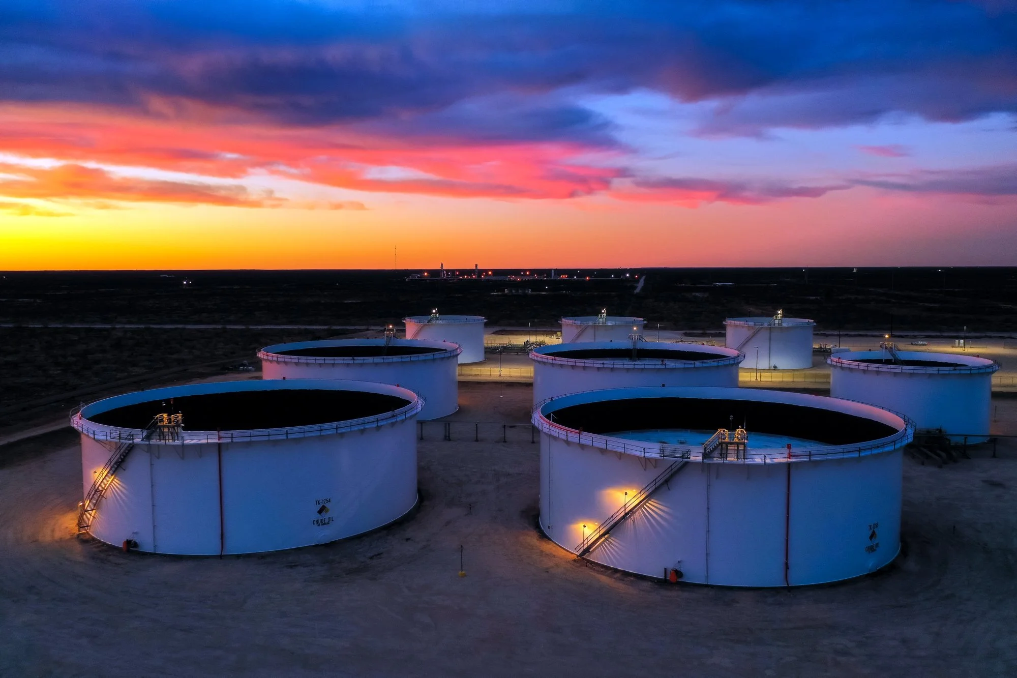 Six large white oil storage tanks at an oil refinery during sunset with colorful clouds in the sky.