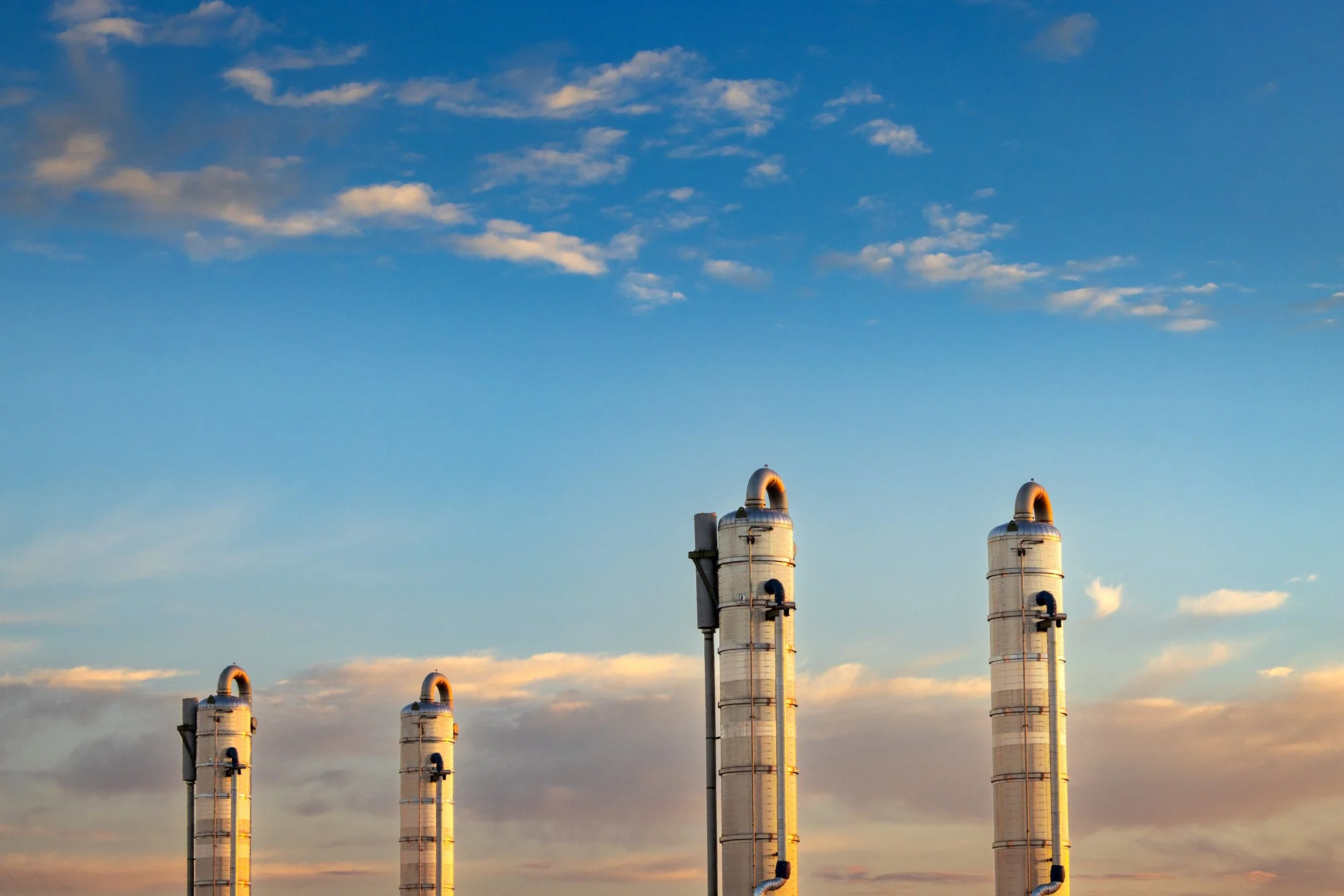 Four large industrial smokestacks against a sky with clouds during sunset.