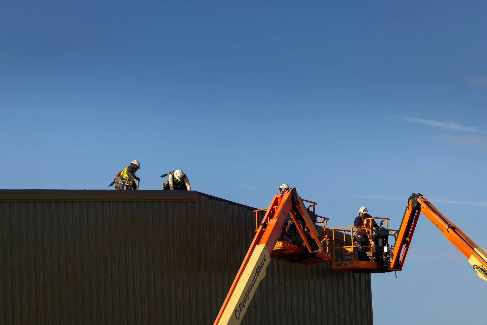 Construction workers in helmets and safety gear working on the roof of a building using a cherry picker lift.