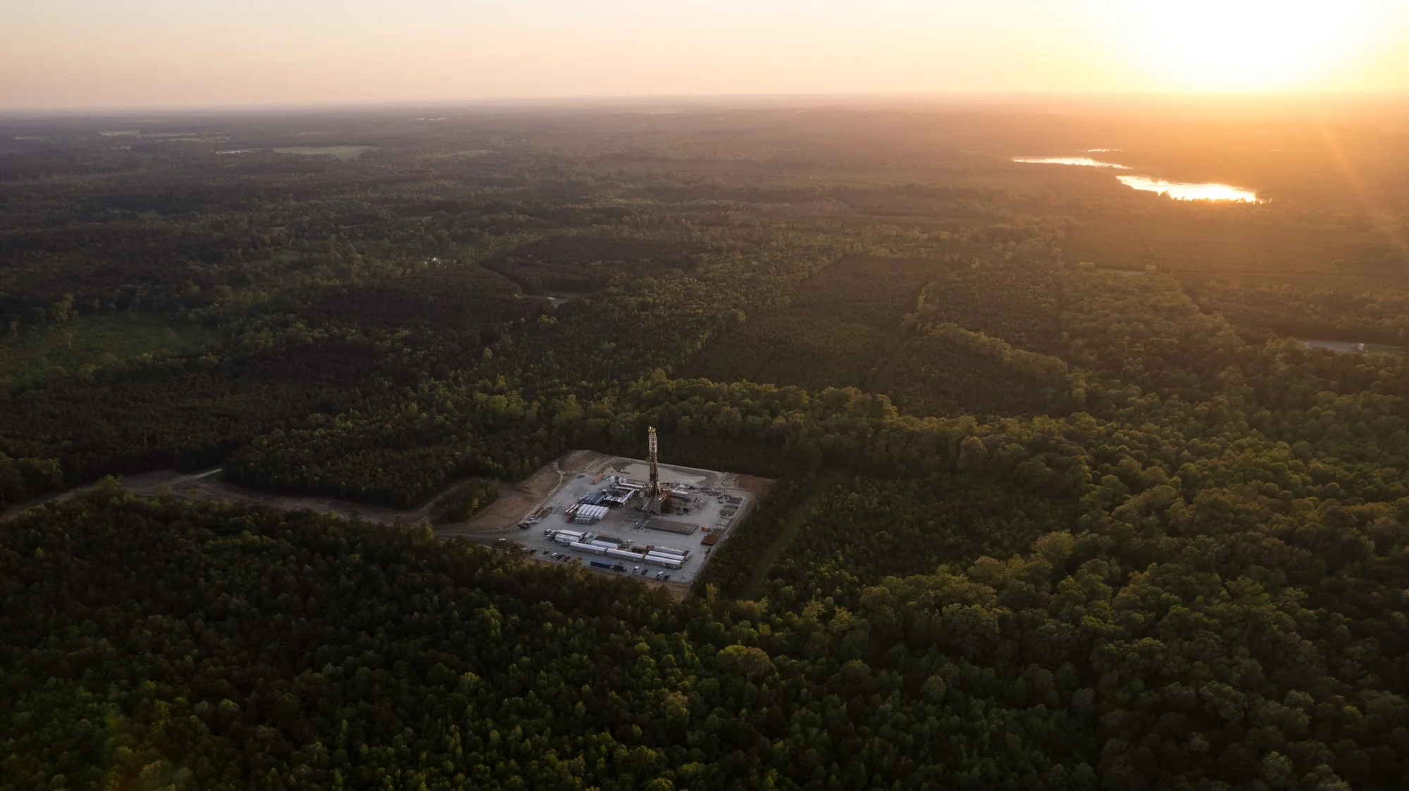 An aerial view of a forested landscape at sunset, with a construction site or industrial facility with a tall crane in the foreground.