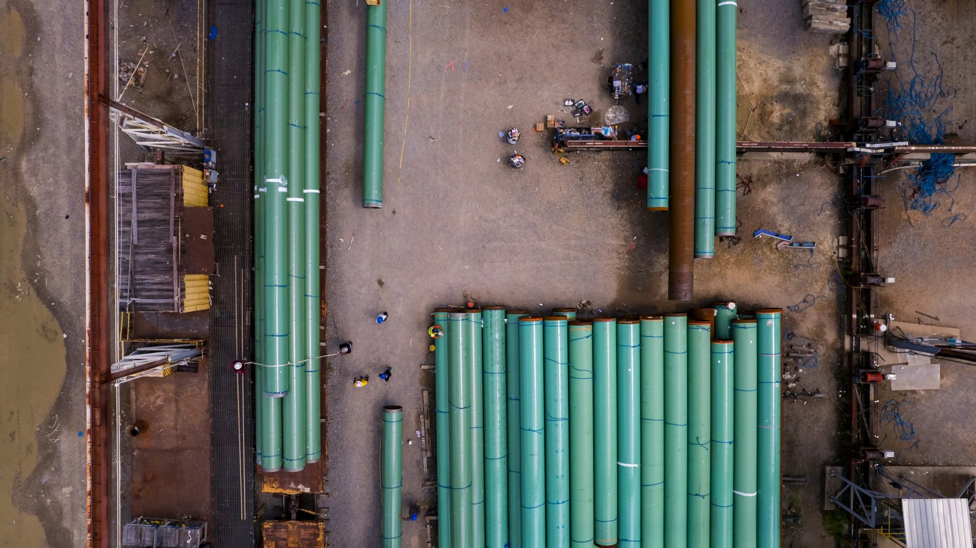 An aerial view of a construction site showing large green pipes arranged in rows, construction workers, and equipment.