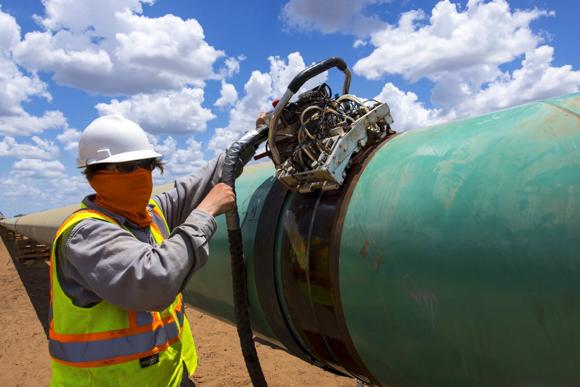 A worker wearing a white hard hat, safety glasses, and an orange face covering, working on a large green pipeline with a mechanical device attached, under a partly cloudy sky.