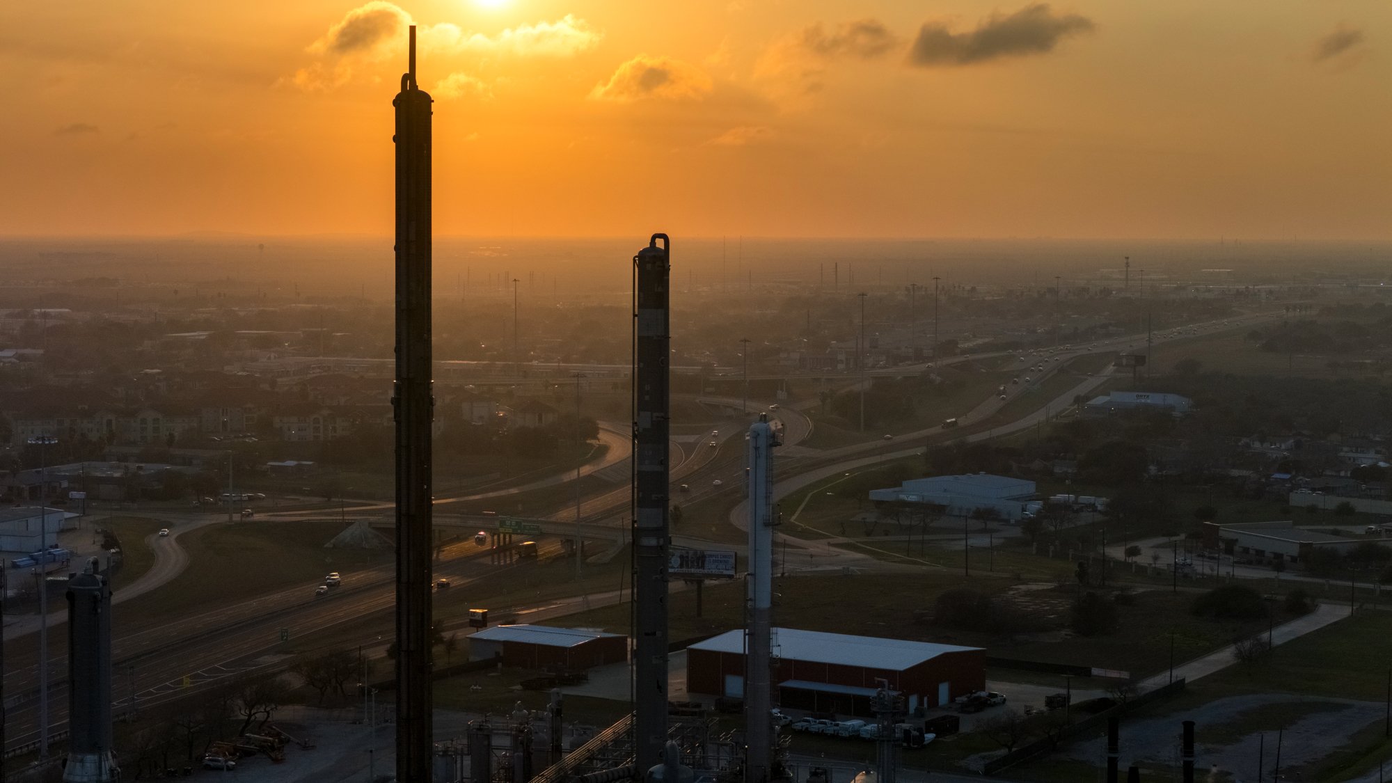 Industrial area with several tall chimneys and a highway, sunset or sunrise creating an orange glow in the sky.