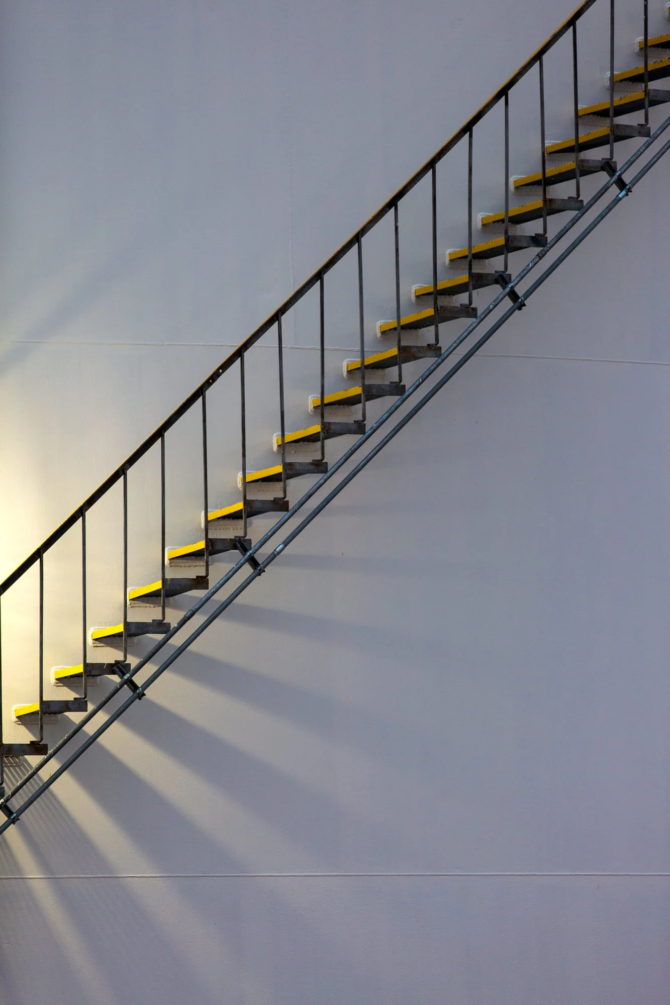 Metal staircase attached to a plain white wall with shadows cast by the steps.
