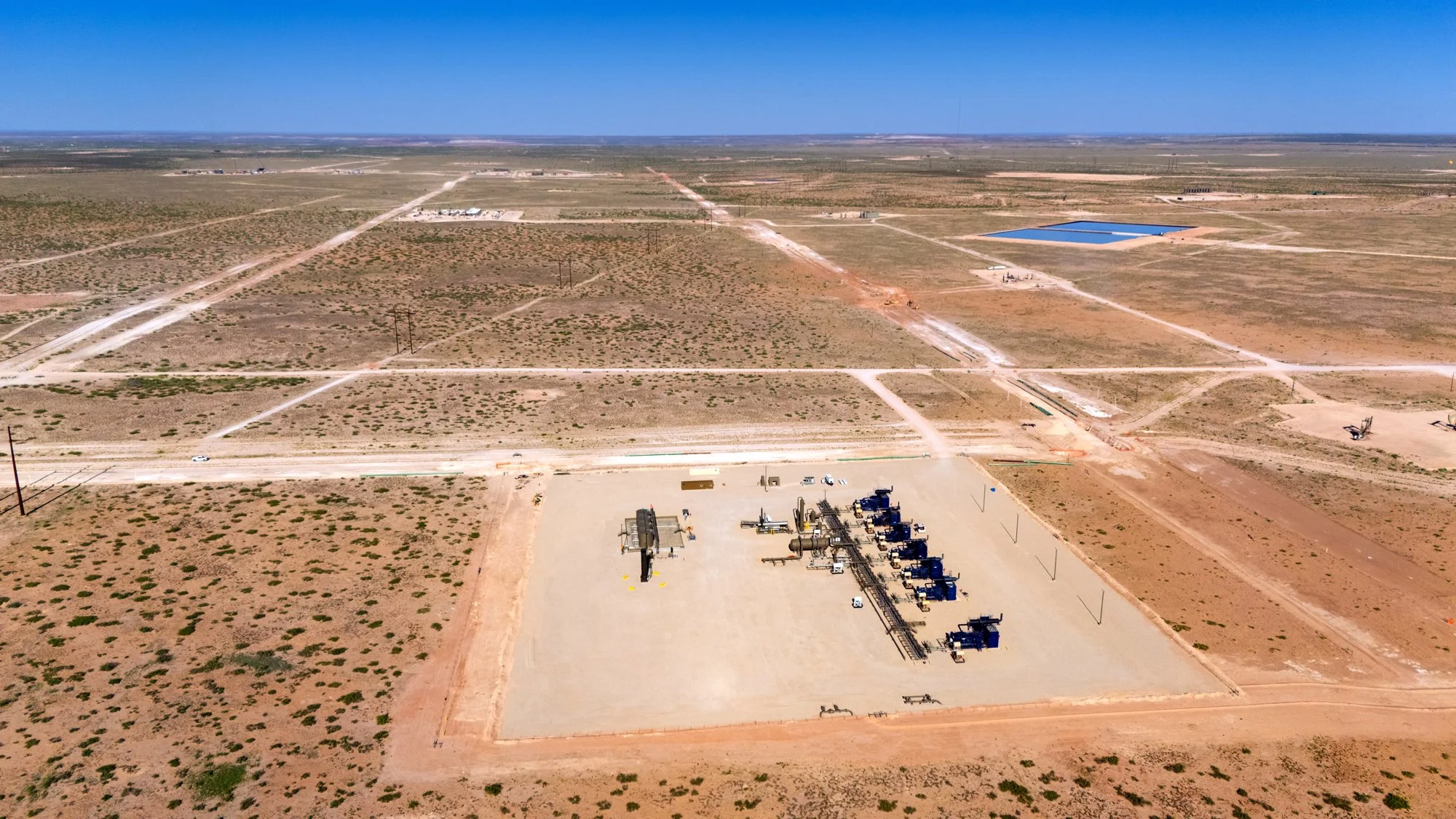 An aerial view of a desert landscape with dirt roads, oil pumping units, and a large water reservoir in the distance.