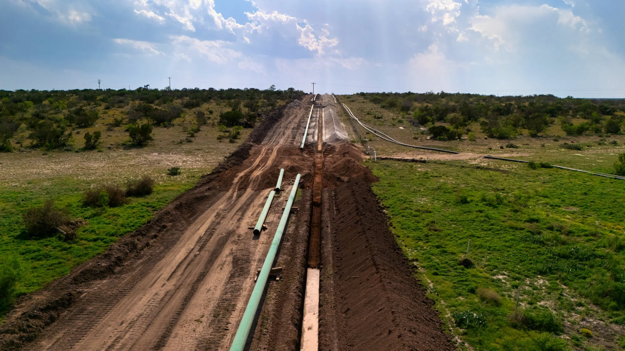 Construction site with large pipes on a dirt road in an open, dry landscape with sparse vegetation under a partly cloudy sky.