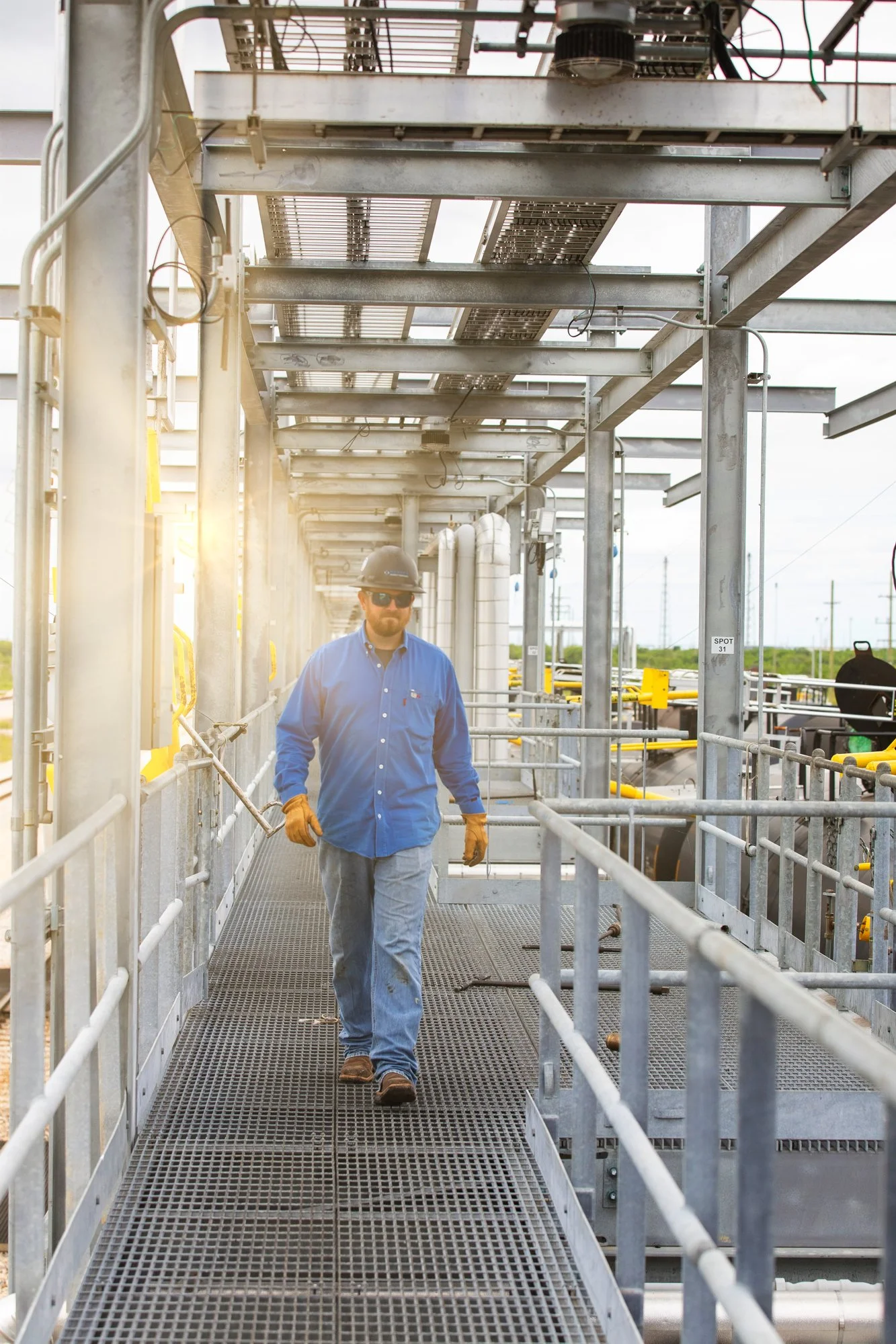 A worker walking on a metal platform at an industrial site with metal scaffolding and piping.