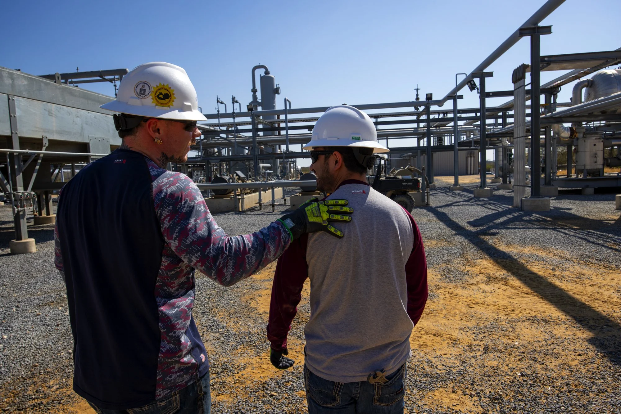 Two workers in safety helmets and gloves stand together in an industrial setting, possibly an oil or chemical plant, with machinery and pipelines in the background.