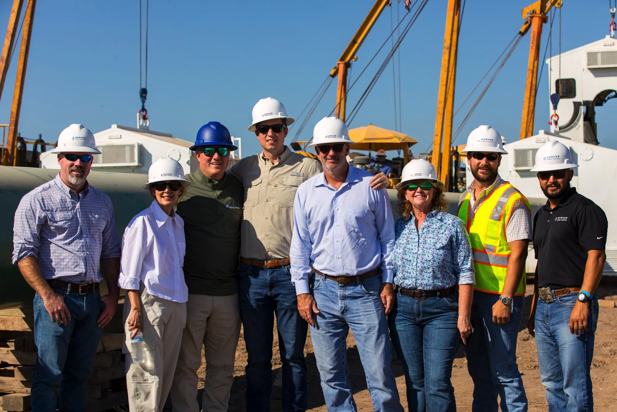 Group of eight construction workers and engineers wearing safety helmets and safety glasses, standing together on a construction site with heavy machinery and cranes in the background.