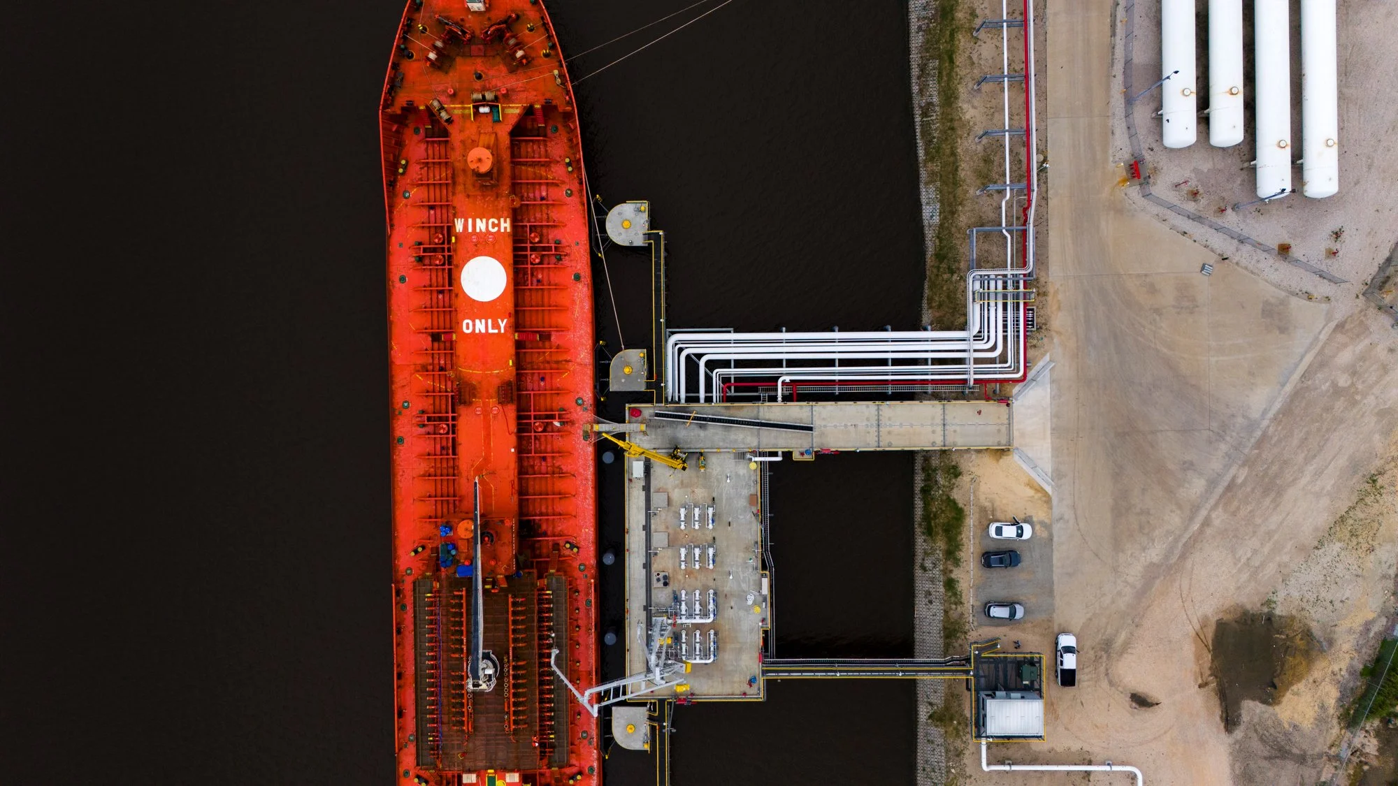 An overhead view of a large orange ship docked at a port, with pipes and industrial structures on land nearby. The ship has the words 'WINCH ONLY' painted on it.