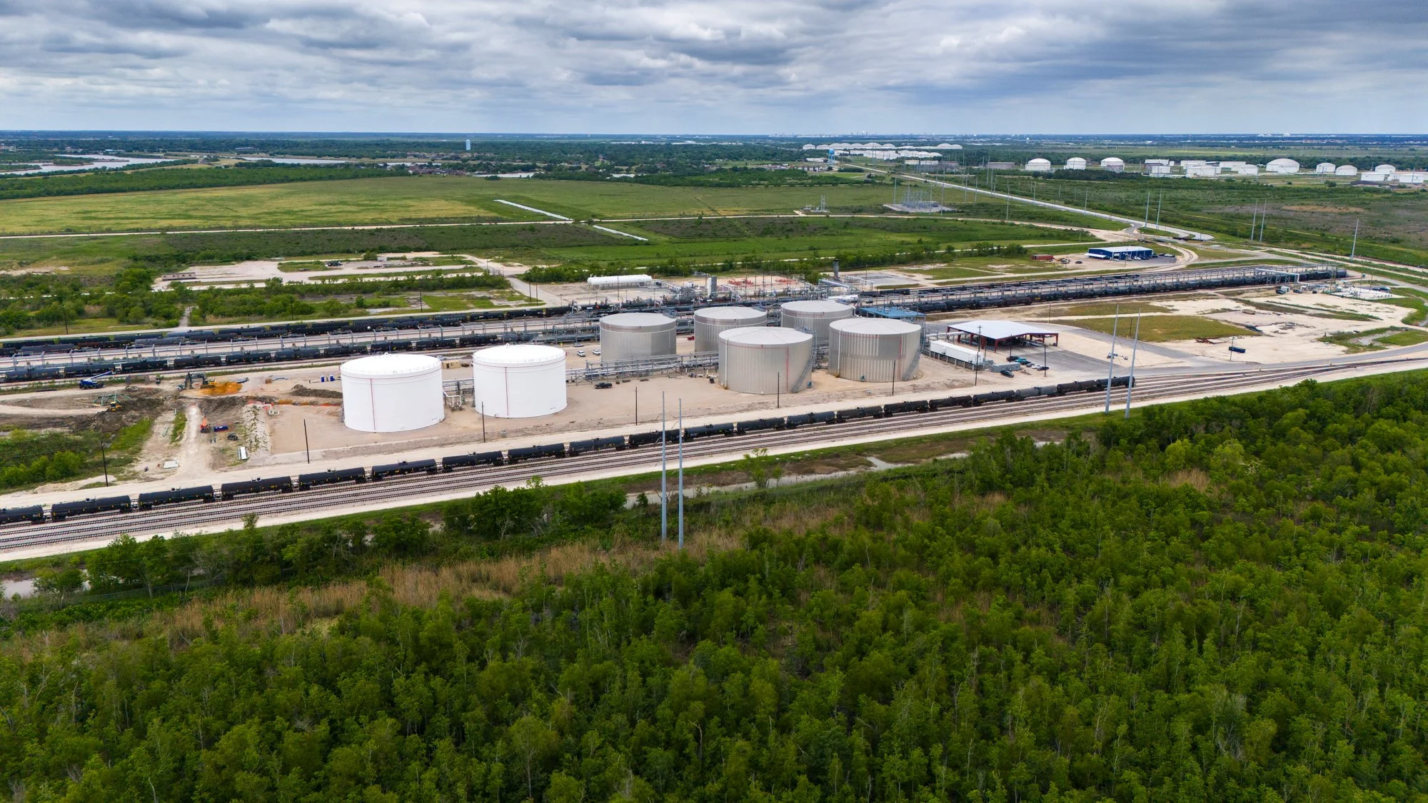 An aerial view of an industrial area with large white storage tanks, railway tracks, and pipelines, surrounded by green trees and open fields under a cloudy sky.