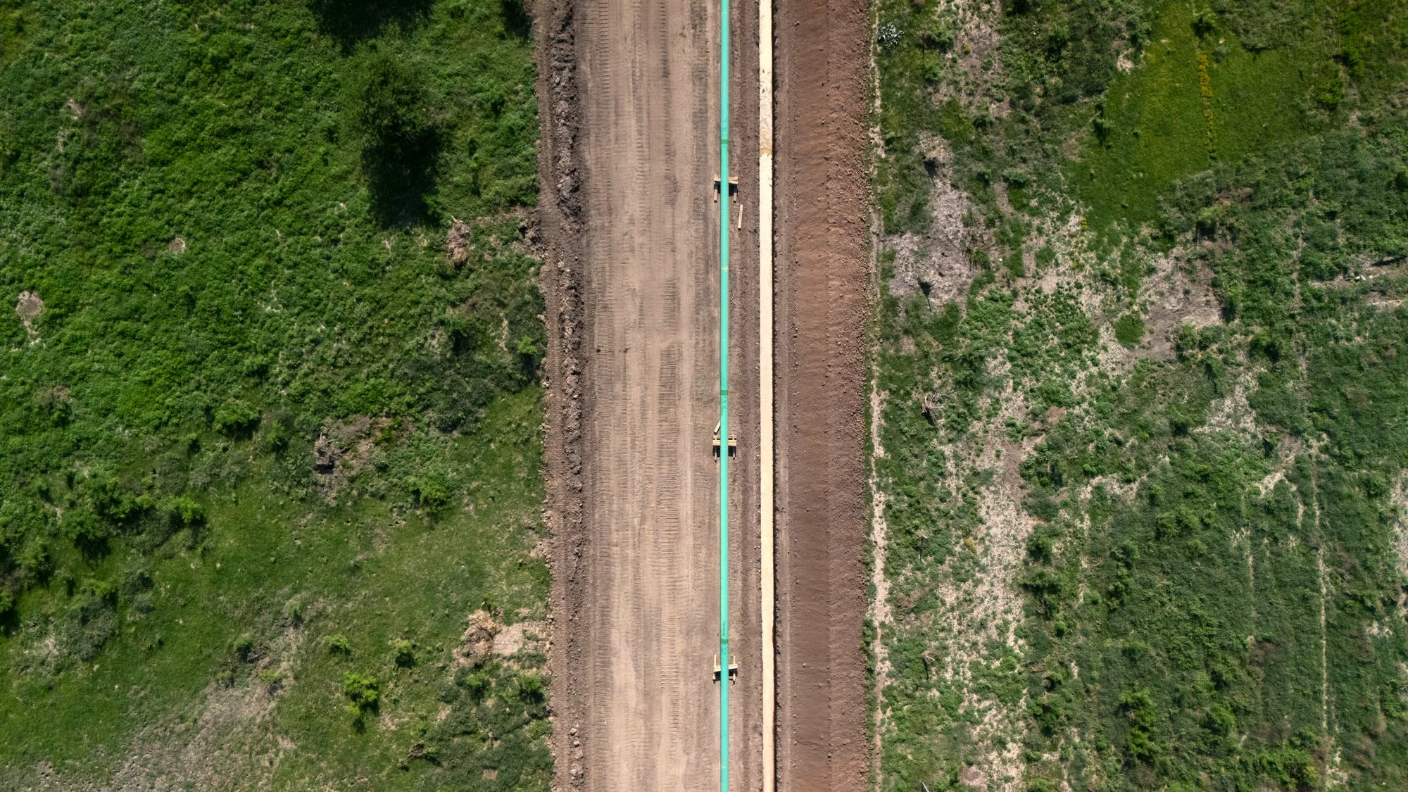 Aerial view of a construction site with a dirt road, green vegetation on both sides, and a large pipeline running in the center.