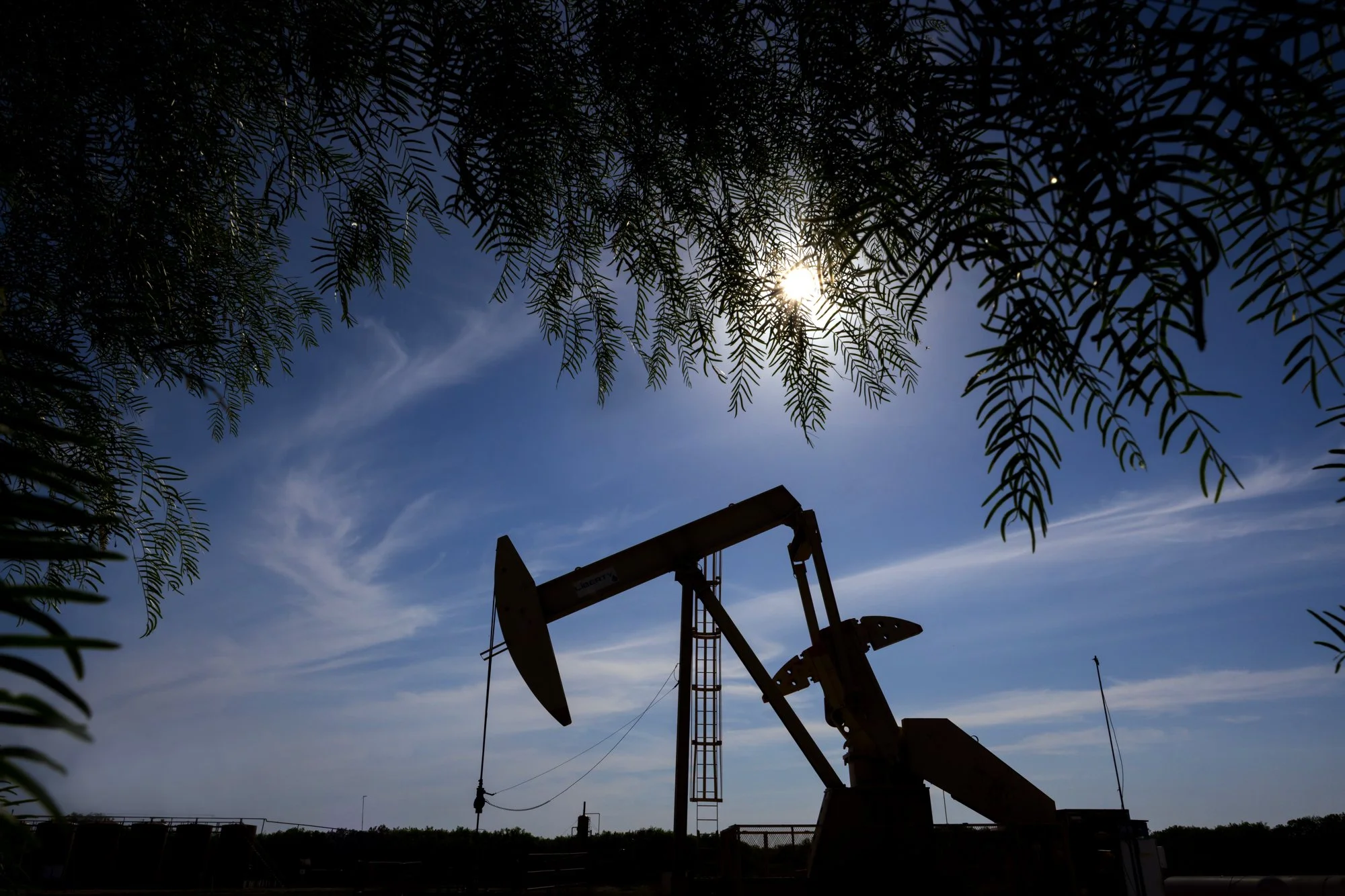 Oil pump jack silhouetted against a bright sky with trees framing the scene.