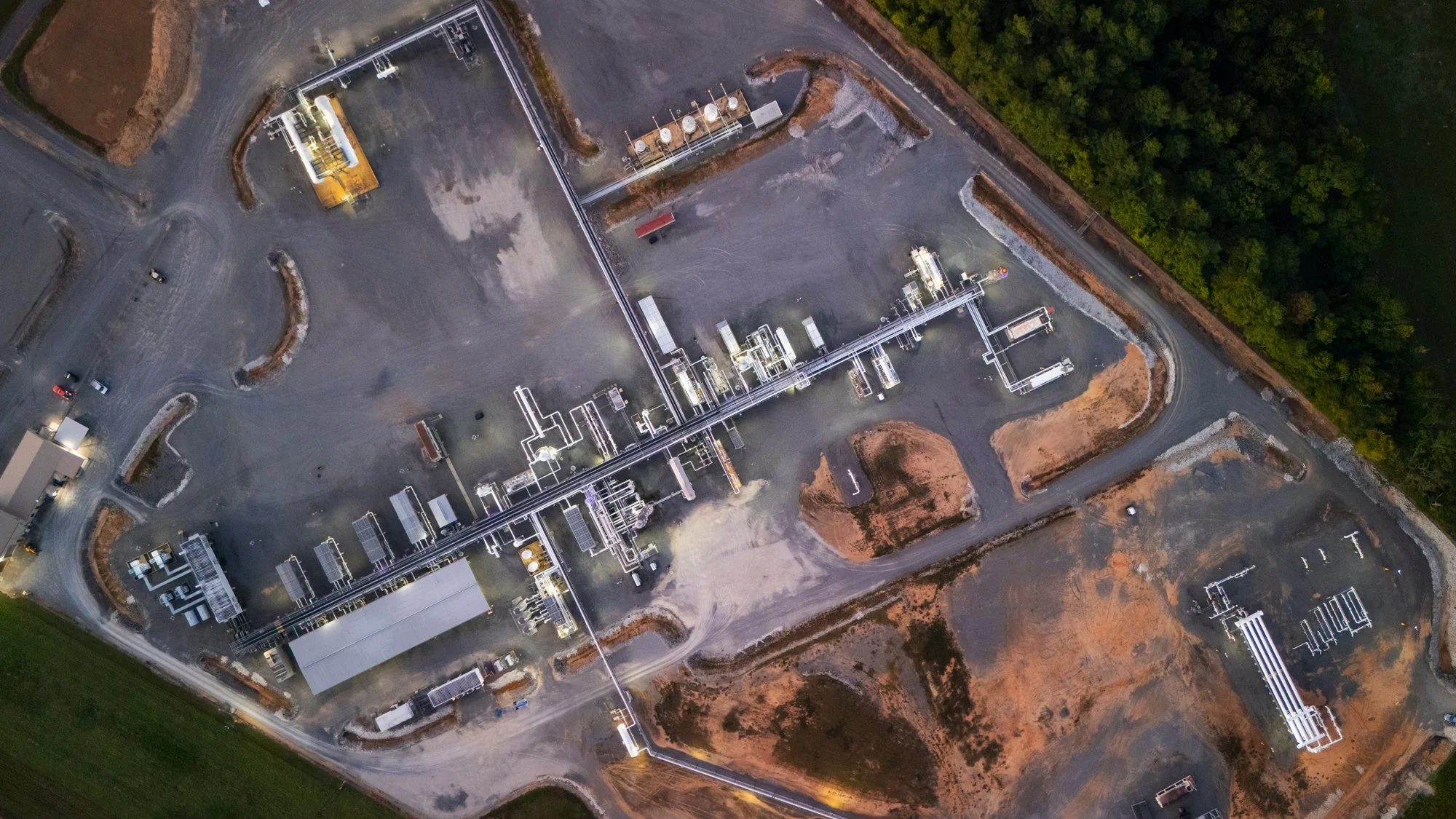An aerial view of an industrial facility with pipelines, equipment, and a parking lot, surrounded by greenery.