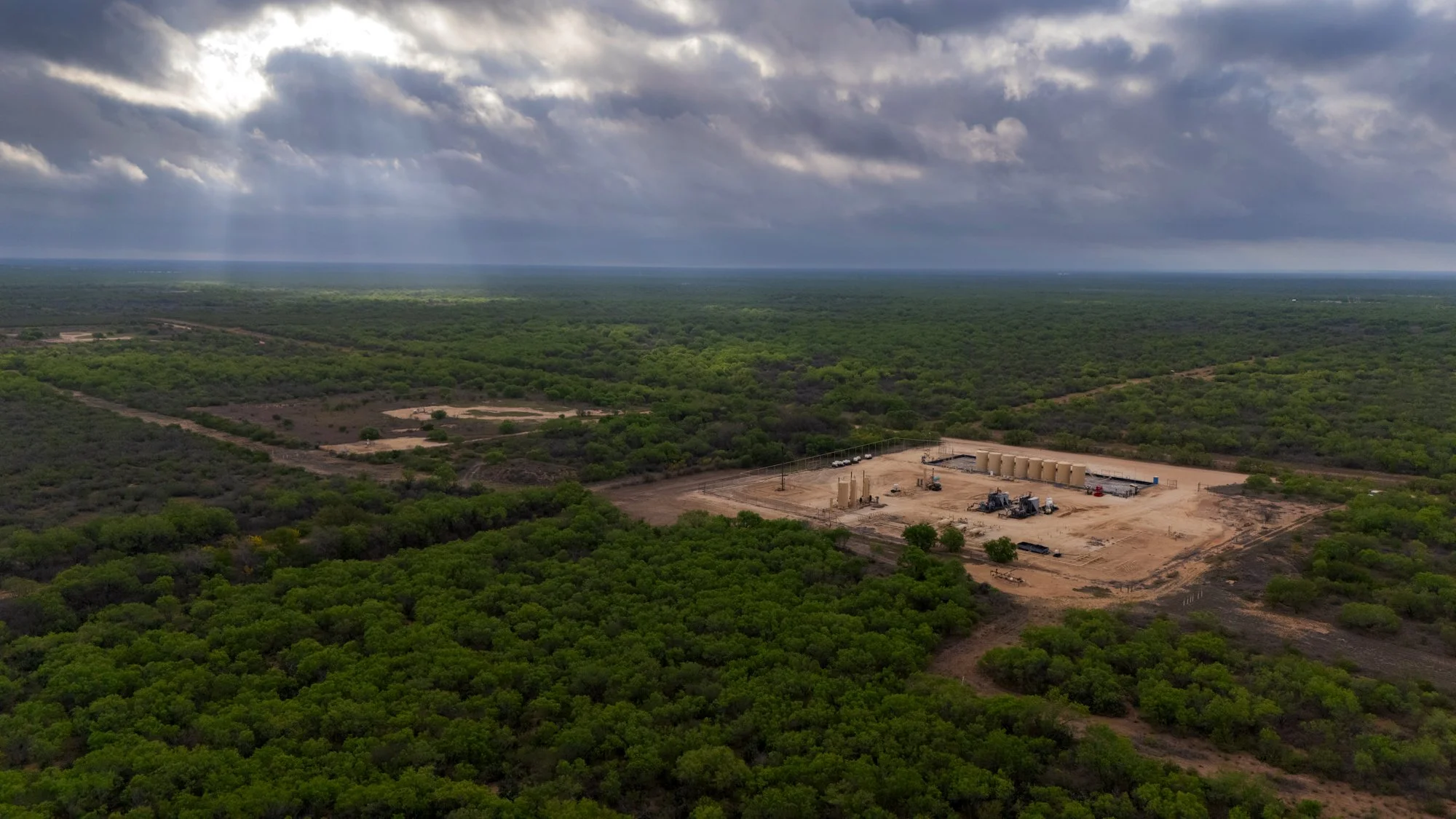 A large cleared construction site with oil or gas tanks and machinery, surrounded by dense green trees, under a cloudy sky.