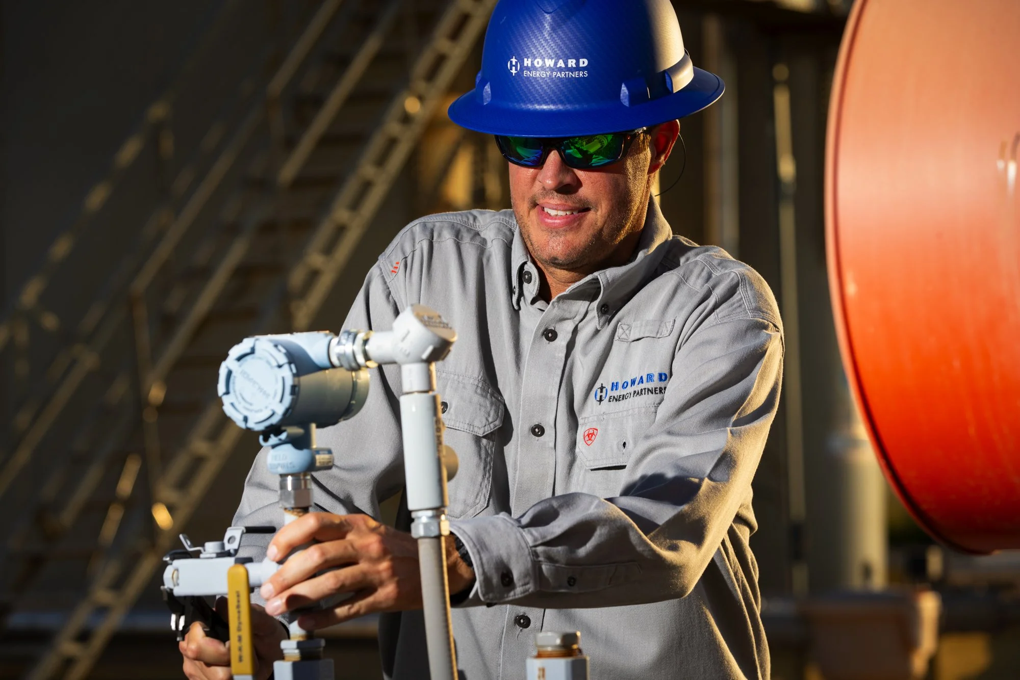 A man wearing safety glasses, a blue hard hat with 'Howard Energy Partners' logo, and a gray shirt working on industrial equipment in an energy facility.