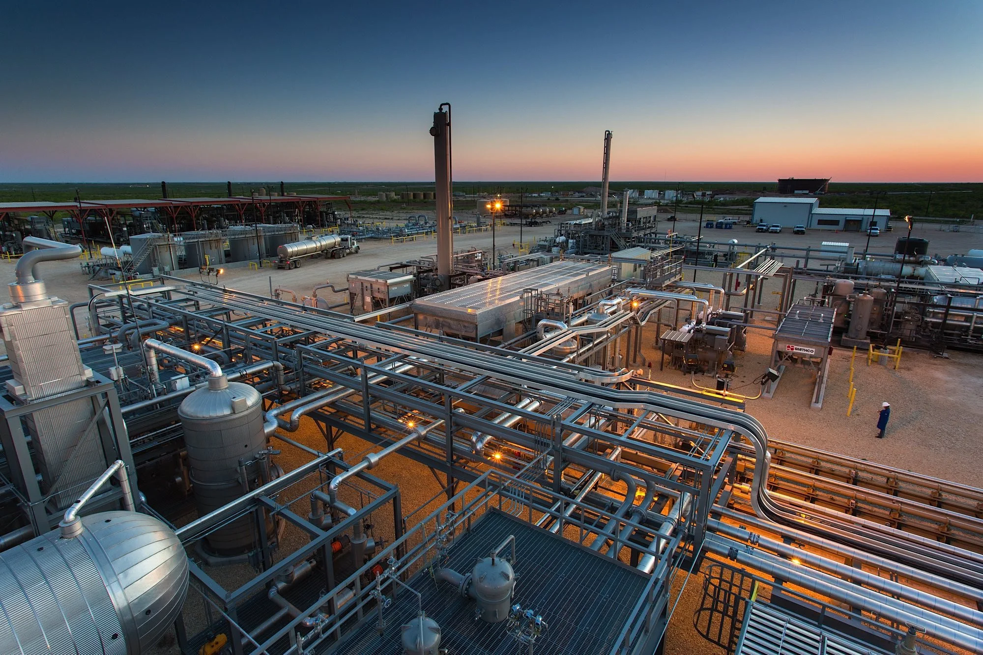 Industrial facility with pipelines, tanks, and equipment at sunset in a desert landscape.
