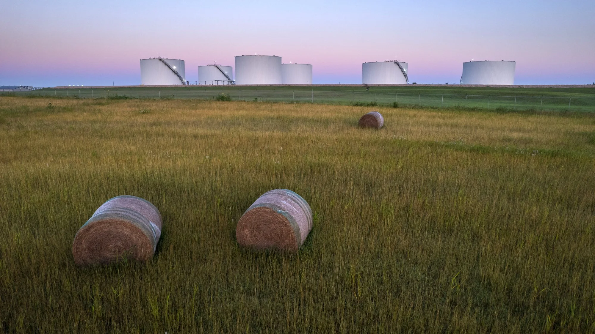 A field with hay bales and large industrial tanks in the background at dusk.