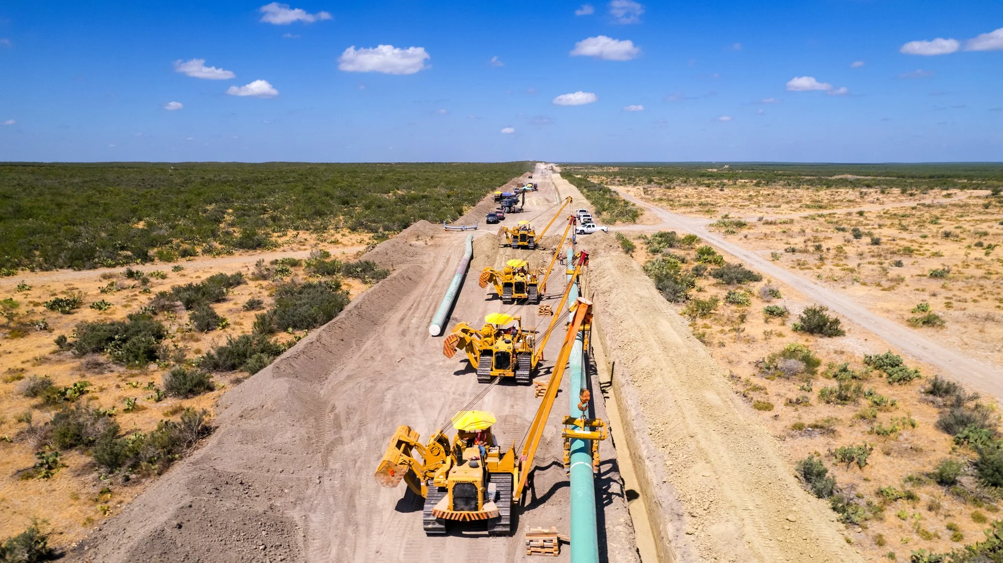 Construction workers and yellow bulldozers working on a pipeline in a desert landscape under a blue sky with scattered clouds.
