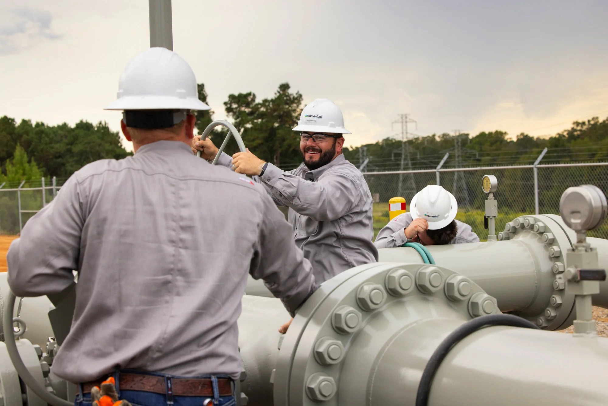 Three workers wearing hard hats and gray shirts working on large industrial pipes outdoors.