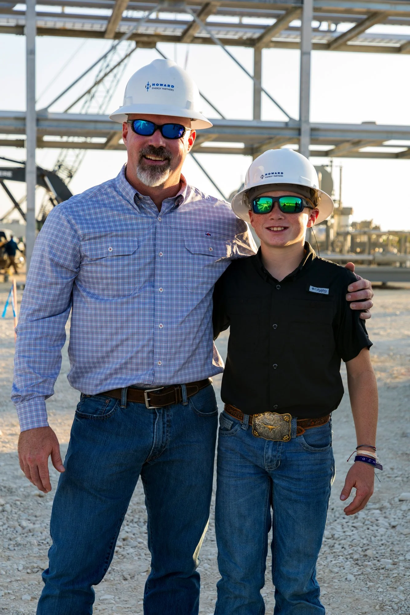 Two smiling men in hard hats and sunglasses standing outdoors at a construction site, with metal framework and equipment in the background.