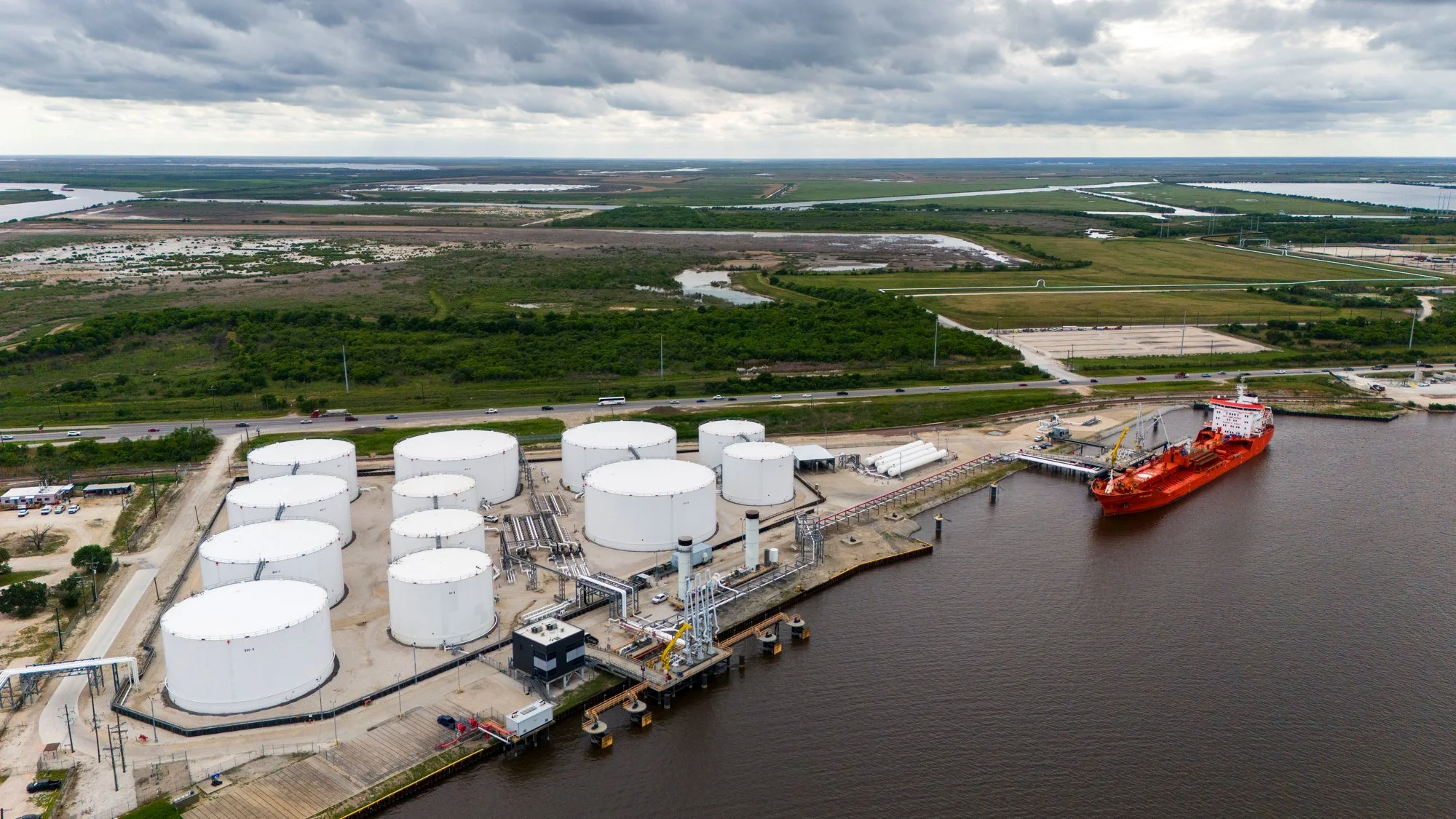 Aerial view of a port facility with large white storage tanks, a cargo ship docked at a pier, and green landscape with wetlands in the background.
