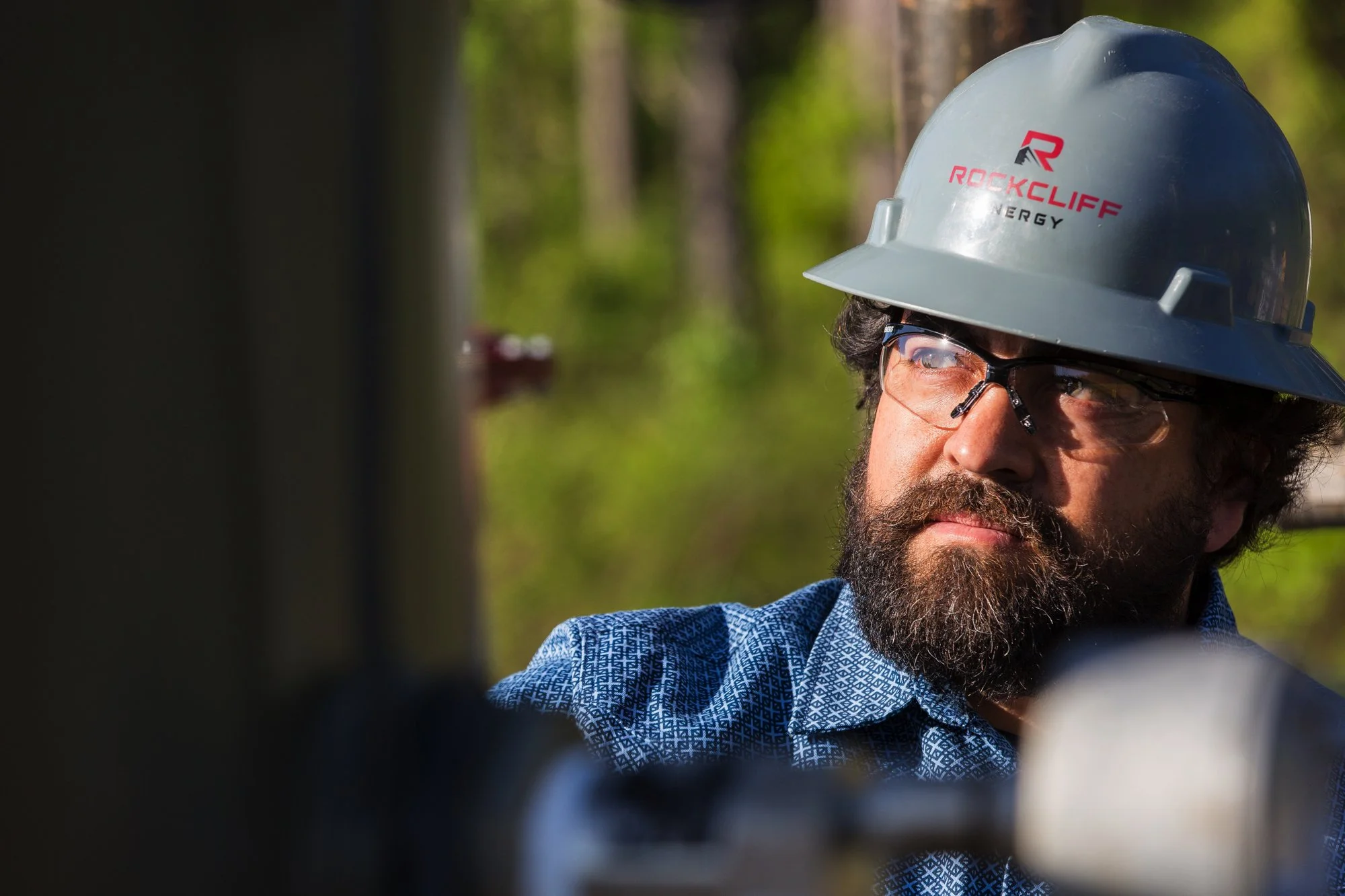 A man with a beard and glasses wearing a gray hard hat and a blue patterned shirt, working outdoors in a wooded area.
