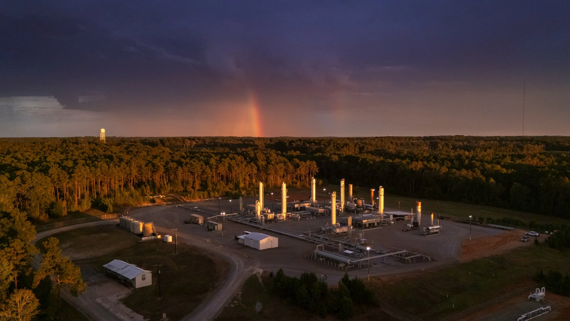 An aerial view of an industrial facility surrounded by a forest with a rainbow in the sky during sunset.
