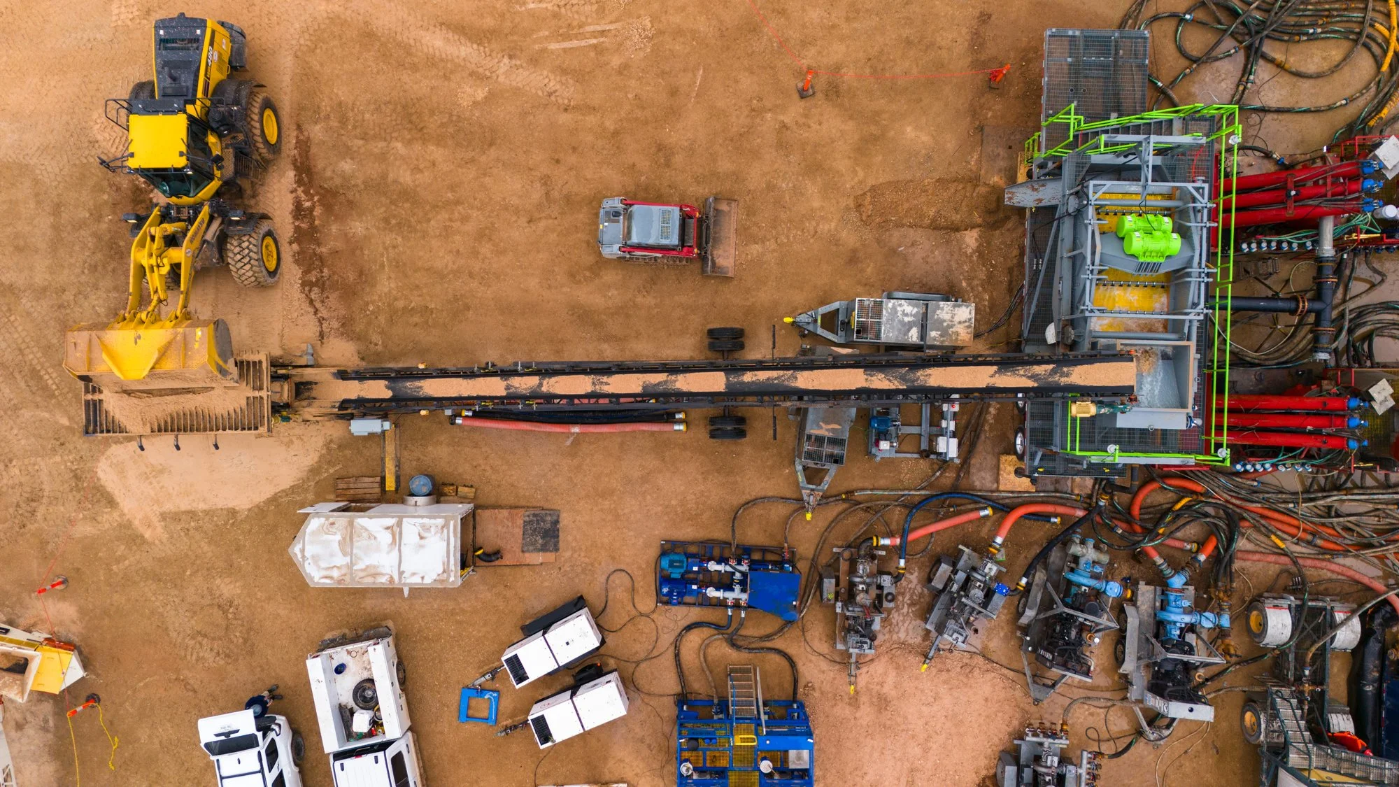 Aerial view of construction site with machinery, equipment, and vehicles on dirt, including a yellow excavator, a snowplow truck, and various pipes and cables.
