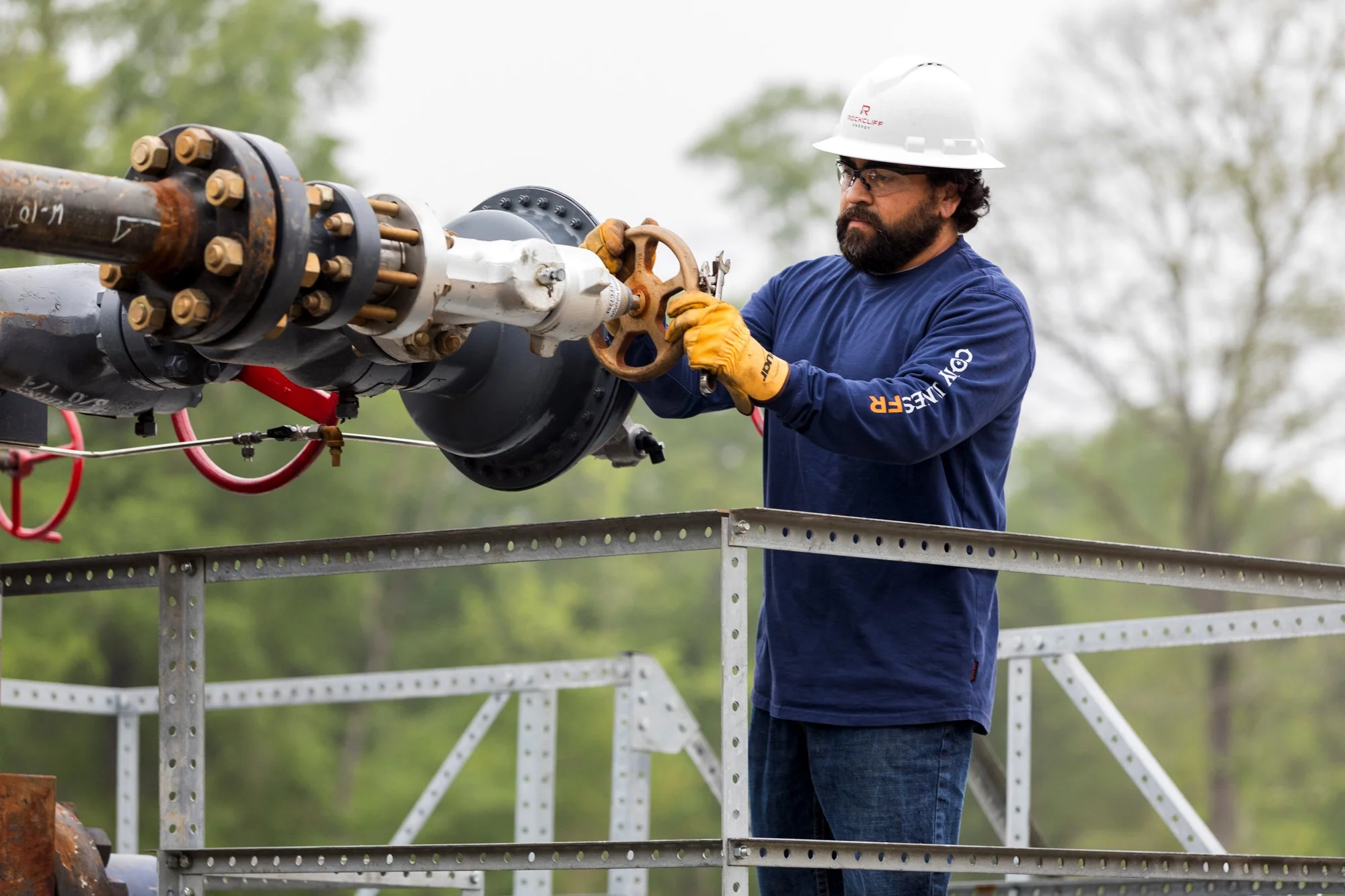 A man wearing safety glasses, a white hard hat, and yellow gloves operates a large valve on an industrial pipeline outdoors, surrounded by a green landscape and overcast sky.