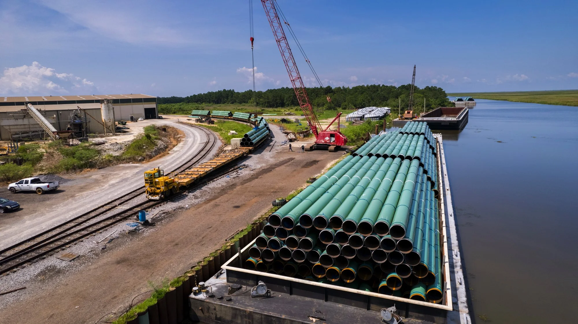 Construction site by a waterway with stacked large green pipes, a crane, construction vehicles, and workers.