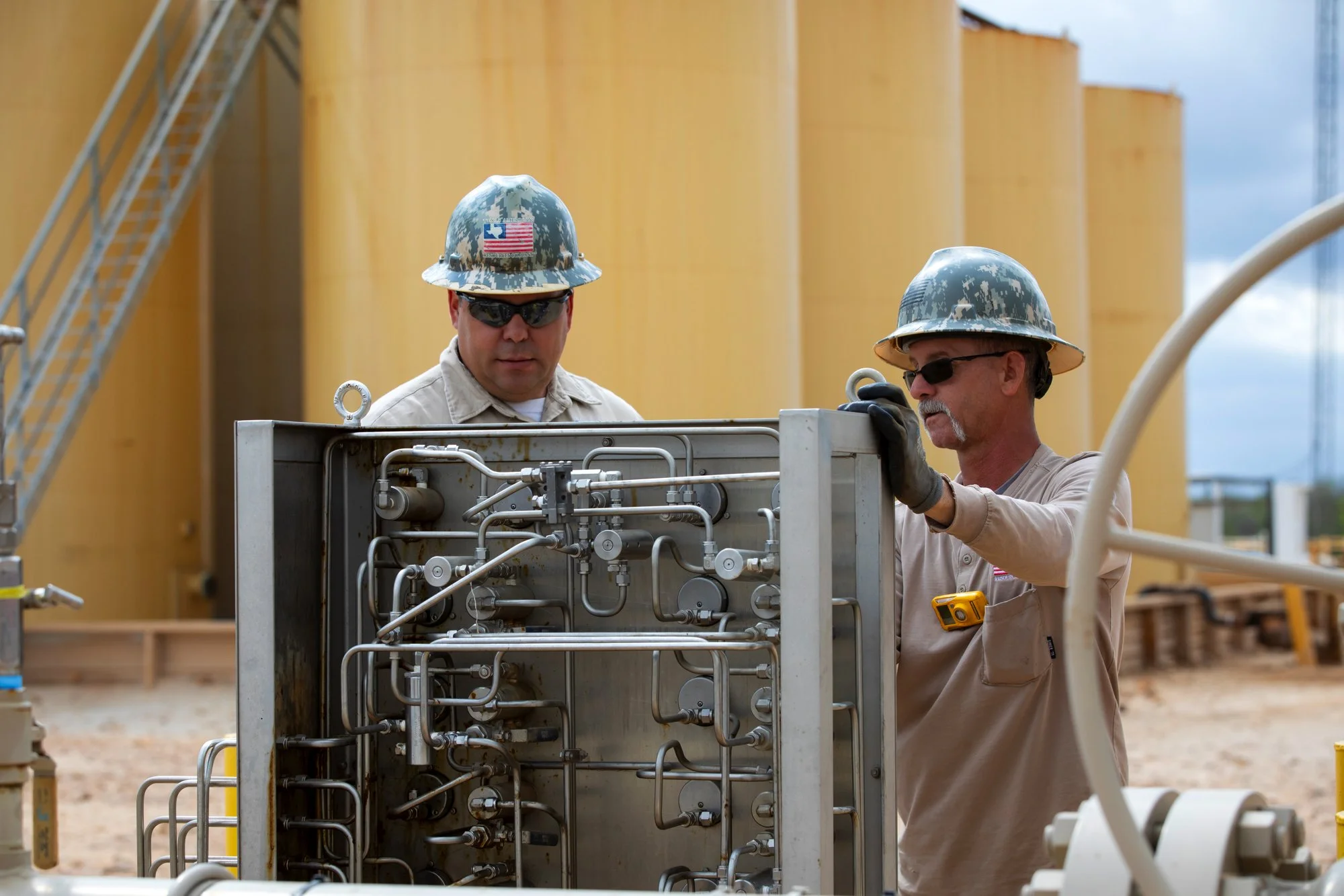 Two men working on a piece of industrial equipment at an outdoor construction site, wearing camo helmets with American flags, sunglasses, and beige work clothes.