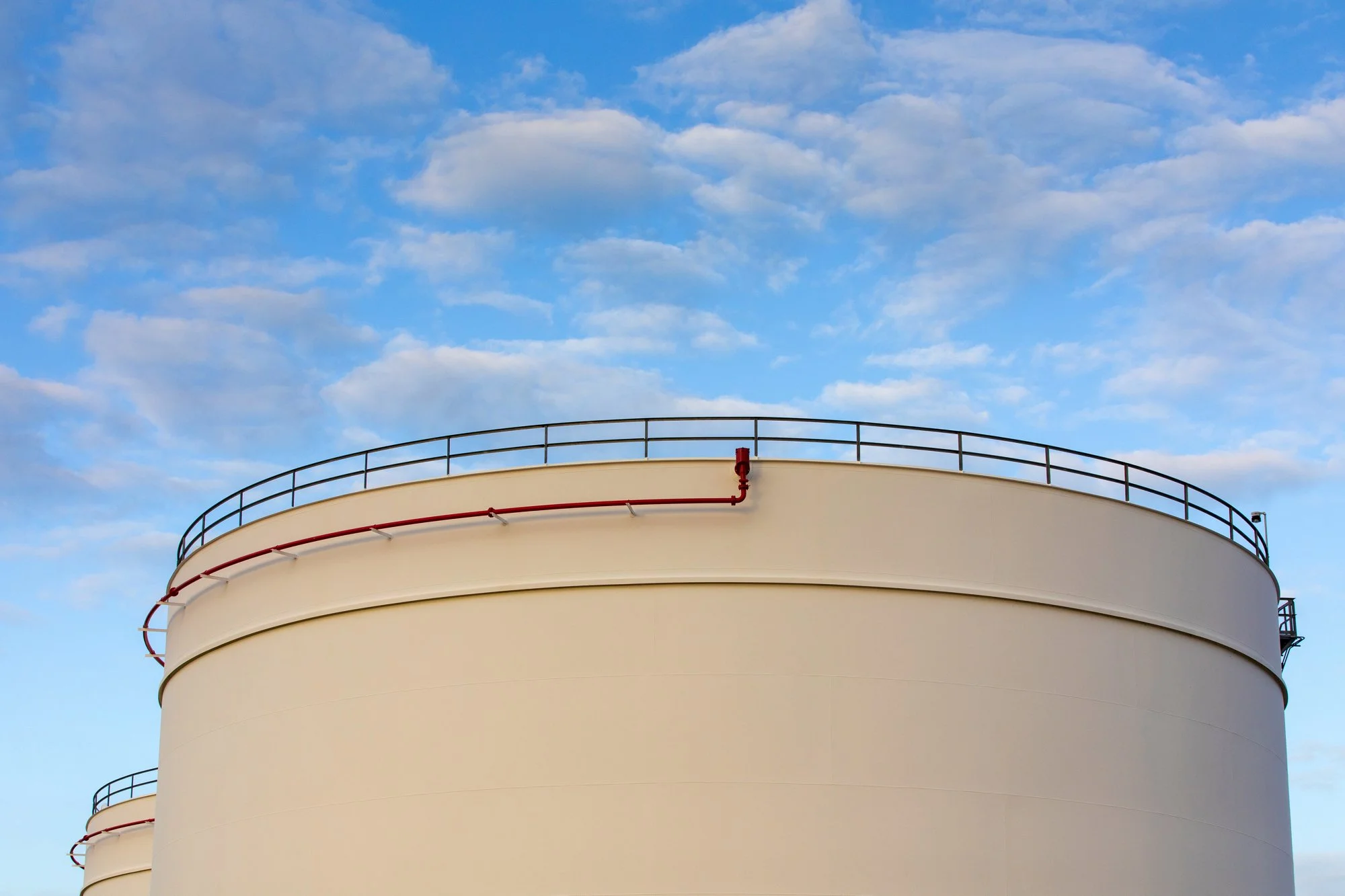 Large white industrial storage tanks with black railings on top, red piping, against a blue sky with scattered white clouds.