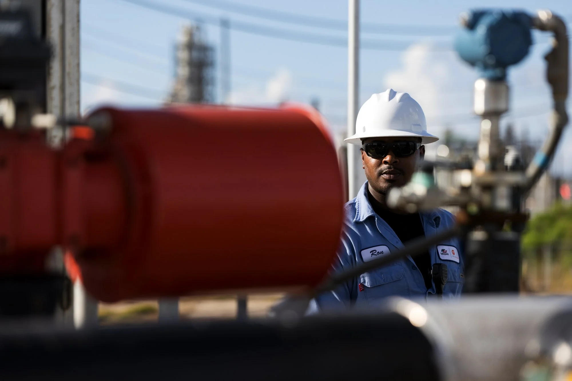 A worker in safety gear, including a white hard hat and sunglasses, looking at industrial equipment at an outdoor facility with power lines and a building in the background.
