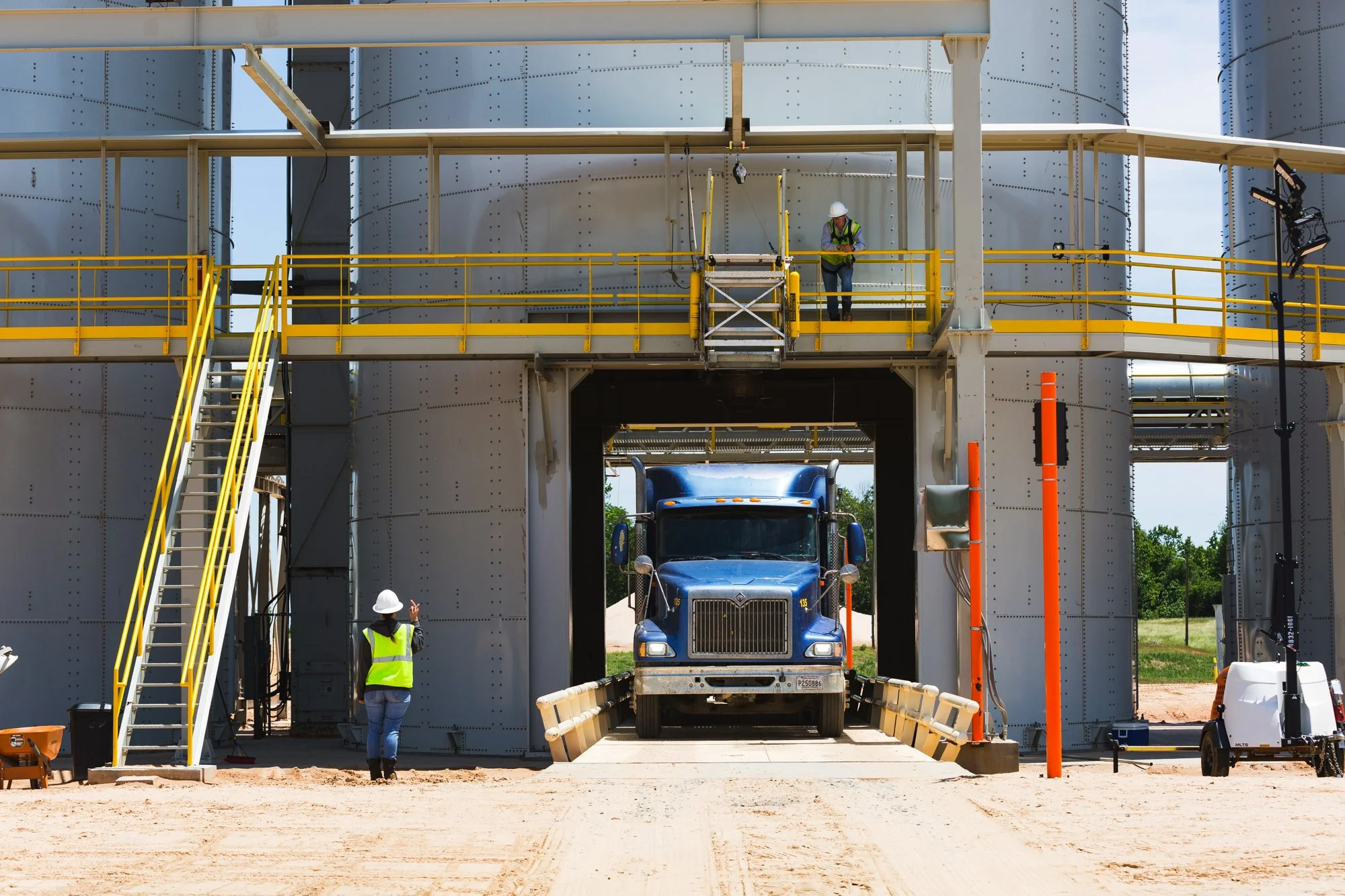 Construction workers in safety vests and helmets working on a large industrial structure with a big blue truck passing through a gap in the structure.
