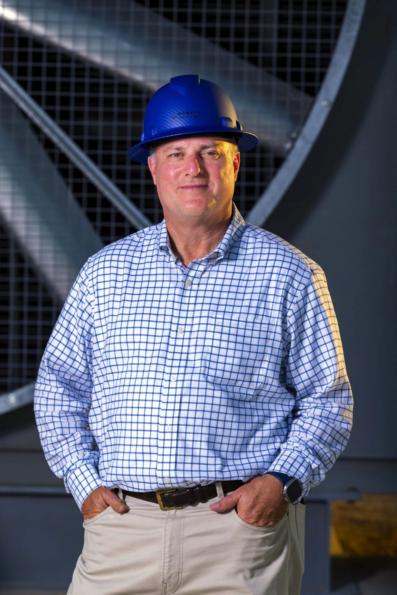 A middle-aged man wearing a blue safety helmet, checkered shirt, and beige pants, standing with hands in pockets in an industrial setting with metal structures in the background.