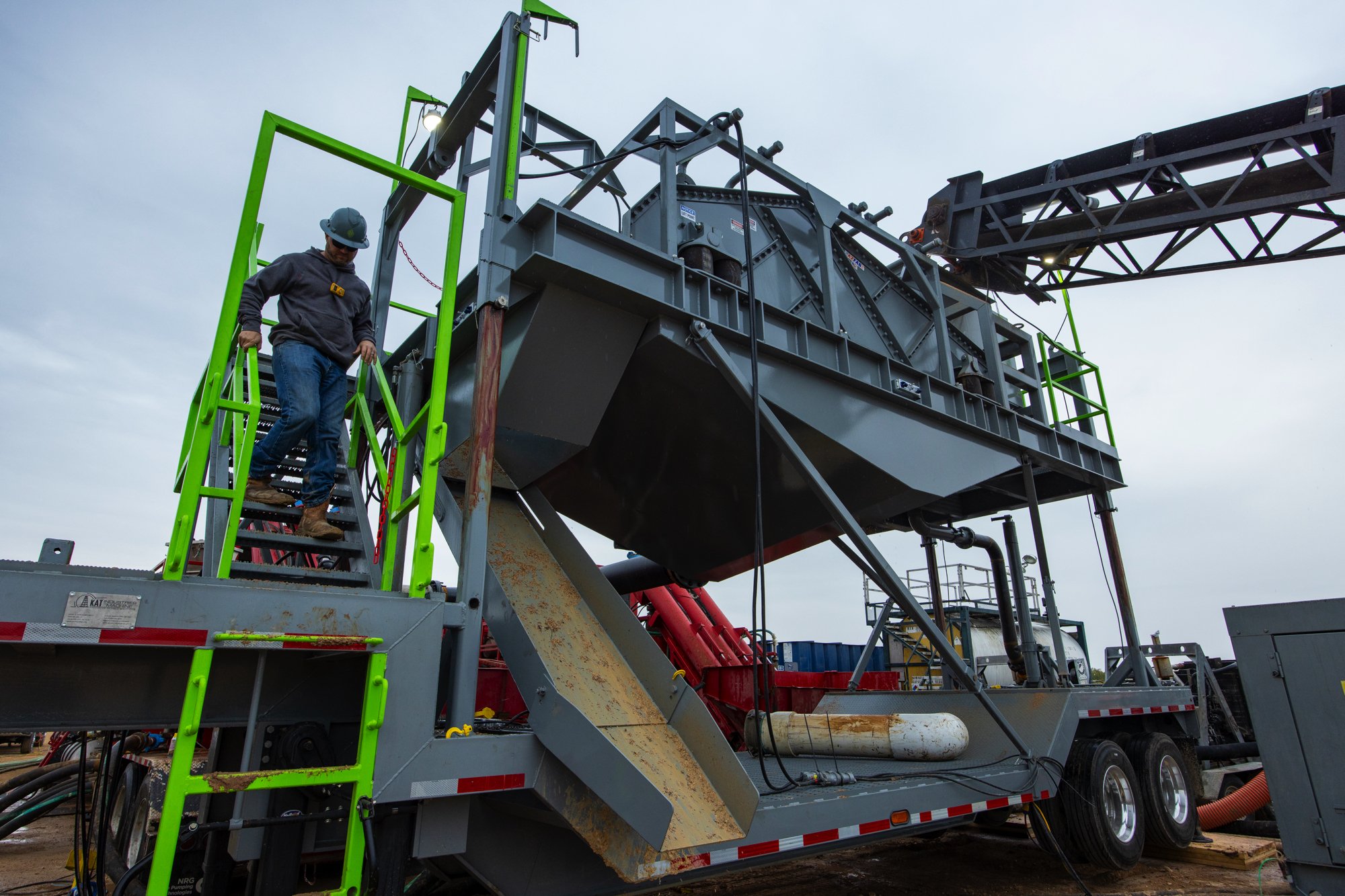 A worker in a gray hoodie, jeans, and a helmet climbs green metal stairs to a large industrial machine mounted on a trailer, with a cloudy sky in the background.