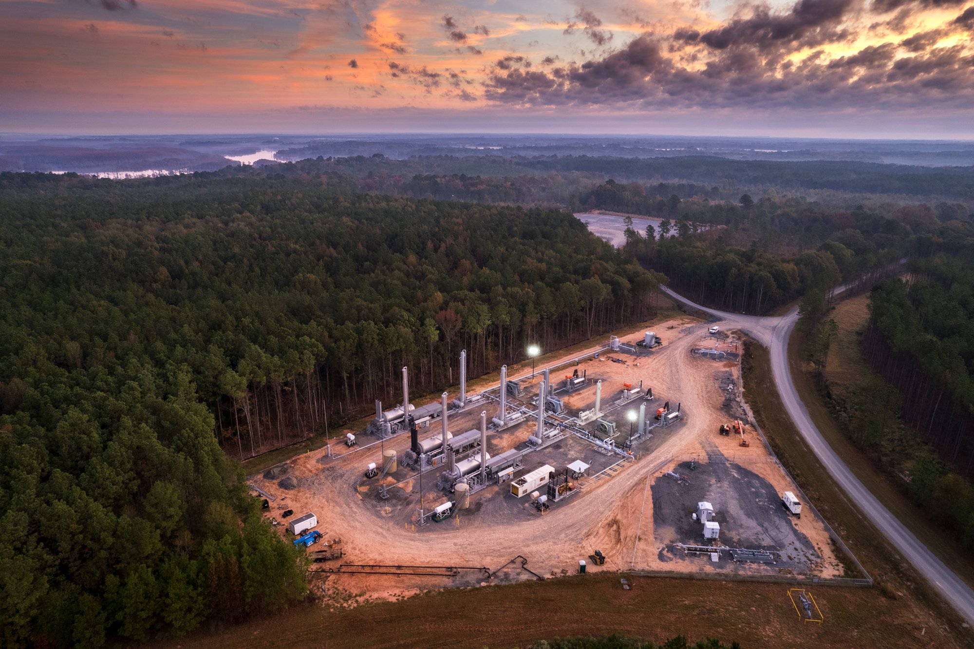 An aerial view of a natural landscape with dense green forests, a winding road, and a small industrial facility or processing plant under construction, at sunset with colorful sky.