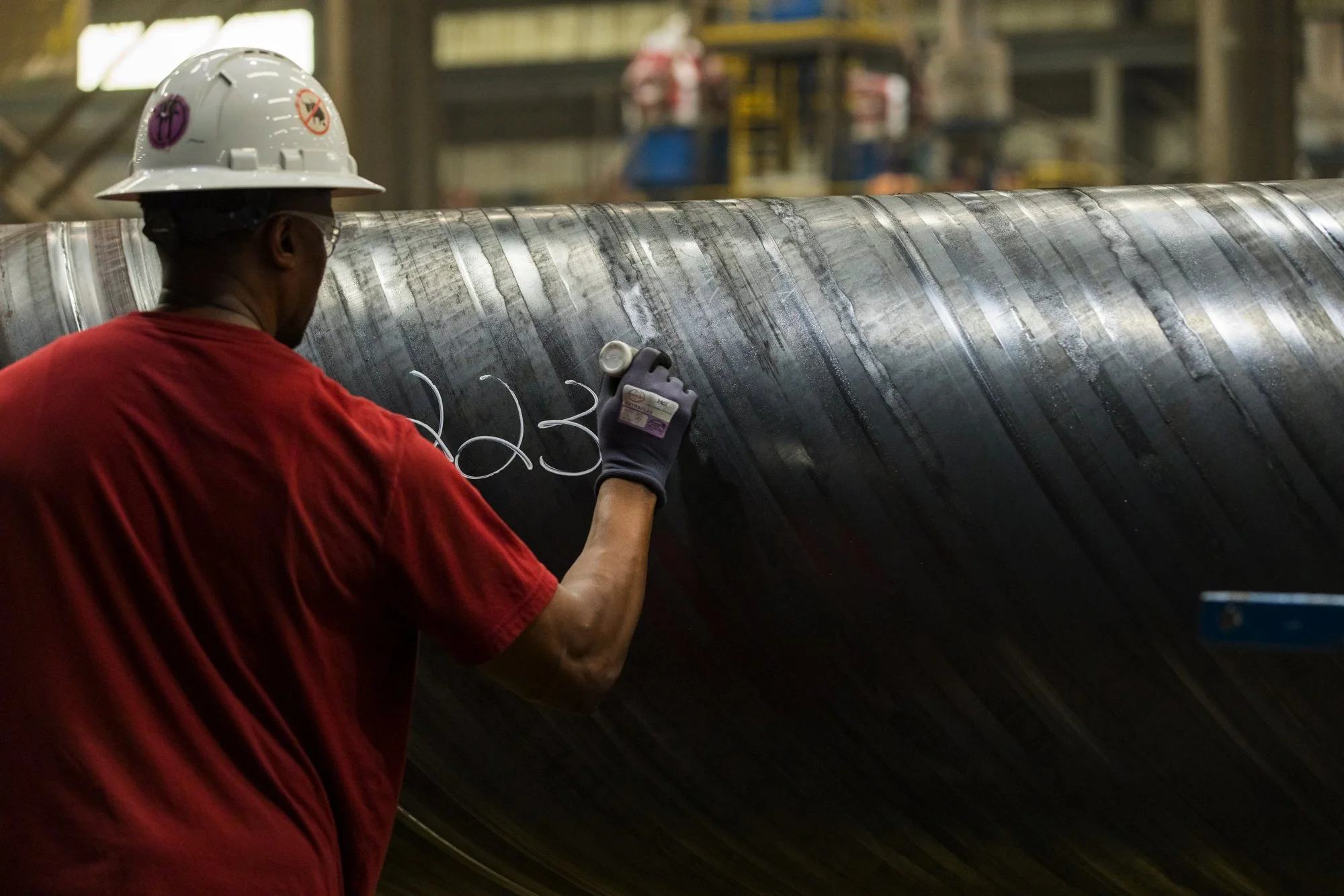 A worker wearing a white safety helmet, glasses, and a red shirt, inspects and writes on a large black industrial pipe in an indoor workshop.